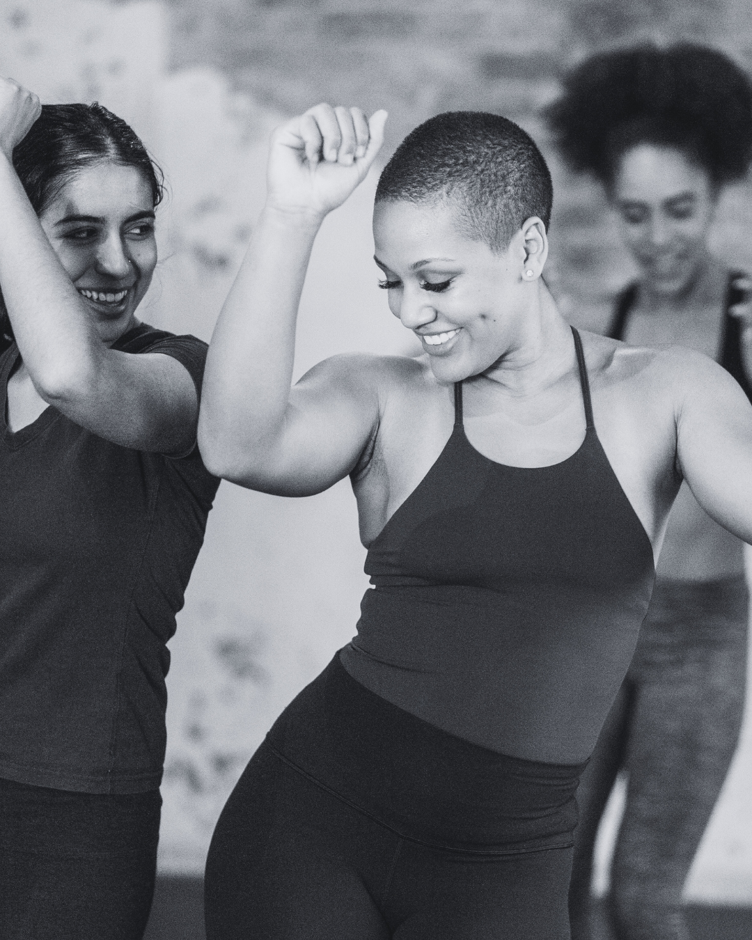 Two women celebrating with smiles, one flexing her arm while the other touches it, in a fitness setting.