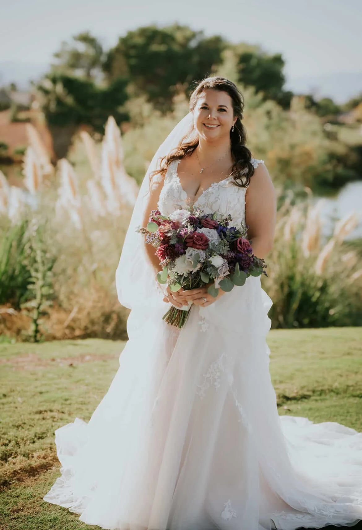 A bride in a white wedding dress holding a bouquet of purple, white, and pink flowers, standing outdoors with trees and a river in the background.