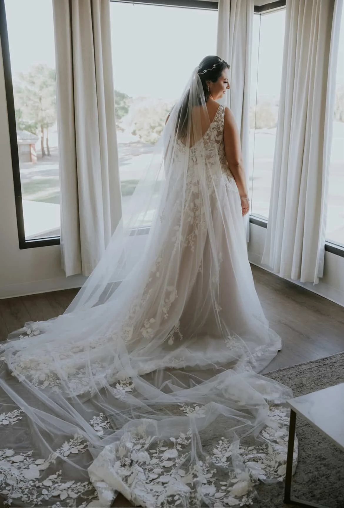 Bride in a white wedding gown and veil standing near large windows with white curtains, looking outside.