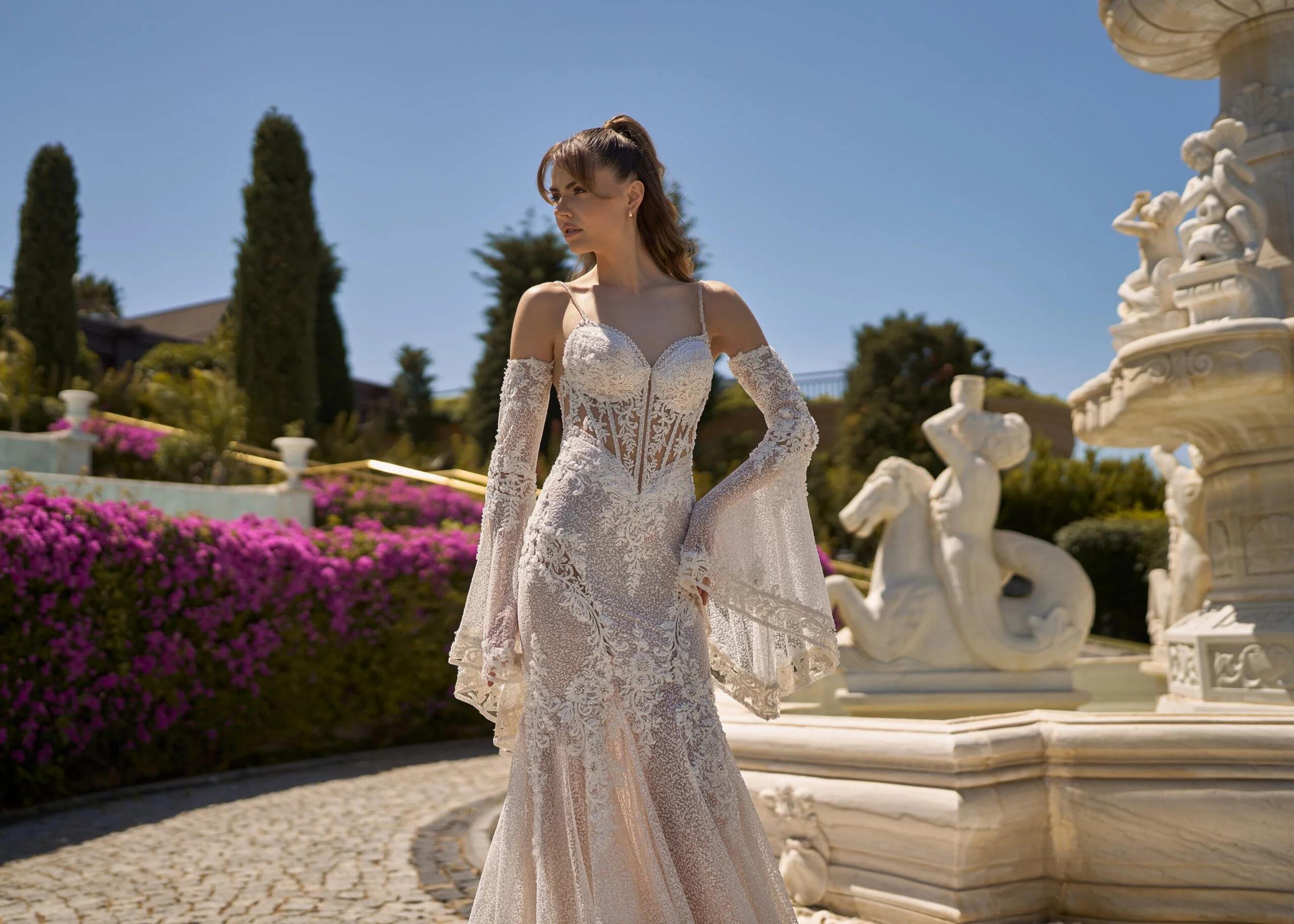 Bride wearing a lace wedding dress with detachable sleeves beside an ornate garden fountain.