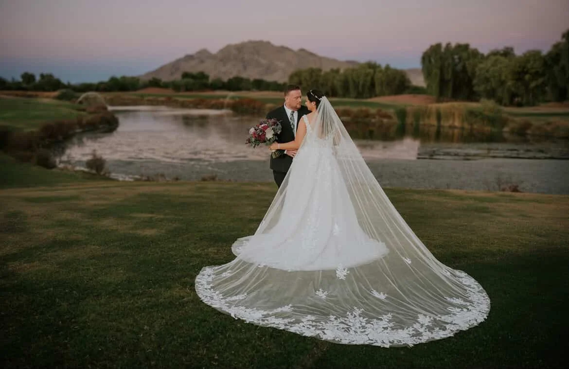 Bride and groom standing on grass by a lake at sunset, with mountains in the background. The bride is wearing a long white wedding gown with a veil, holding a bouquet, and the groom in a dark suit.