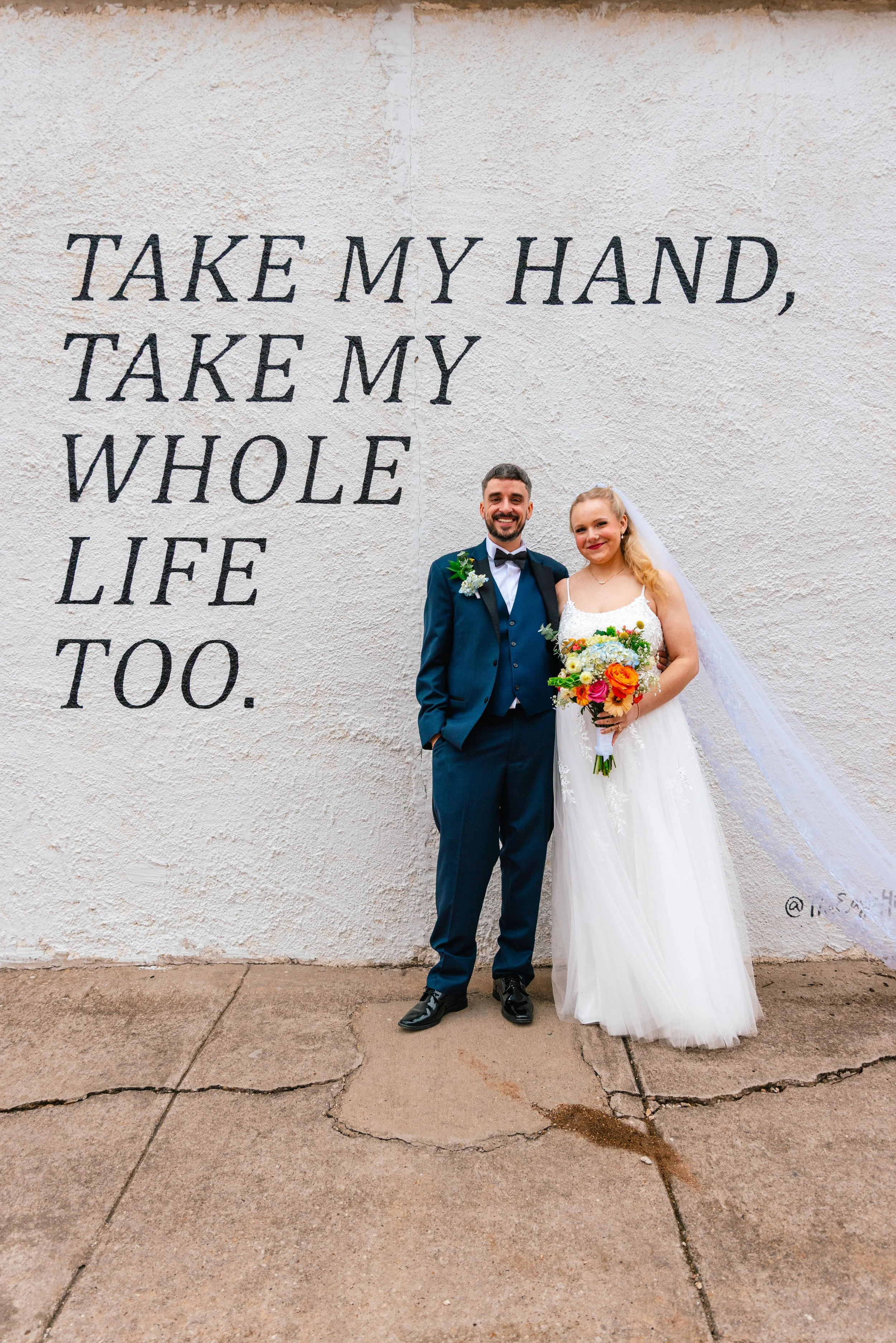 A newlywed couple smiling and standing together in front of a white wall with black text that says "Take my hand, take my whole life too." The groom wears a blue suit with a black bow tie, and the bride wears a white wedding dress holding a colorful bouquet of flowers. The groom has a boutonniere on his suit, and both are smiling at the camera.