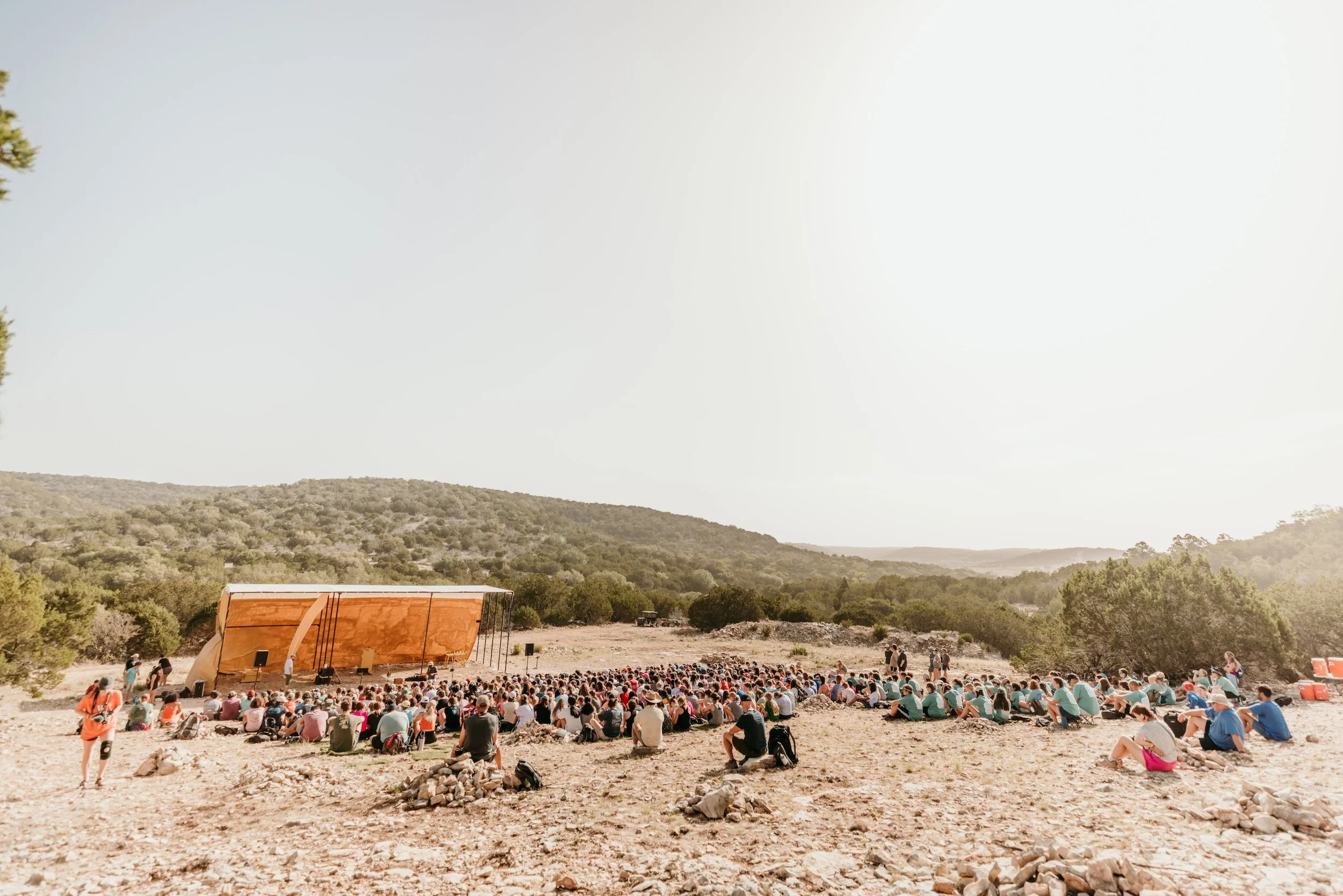 Outdoor concert with a large audience sitting on the ground in a natural setting with hills and trees, a stage with a canopy, and a clear sky.