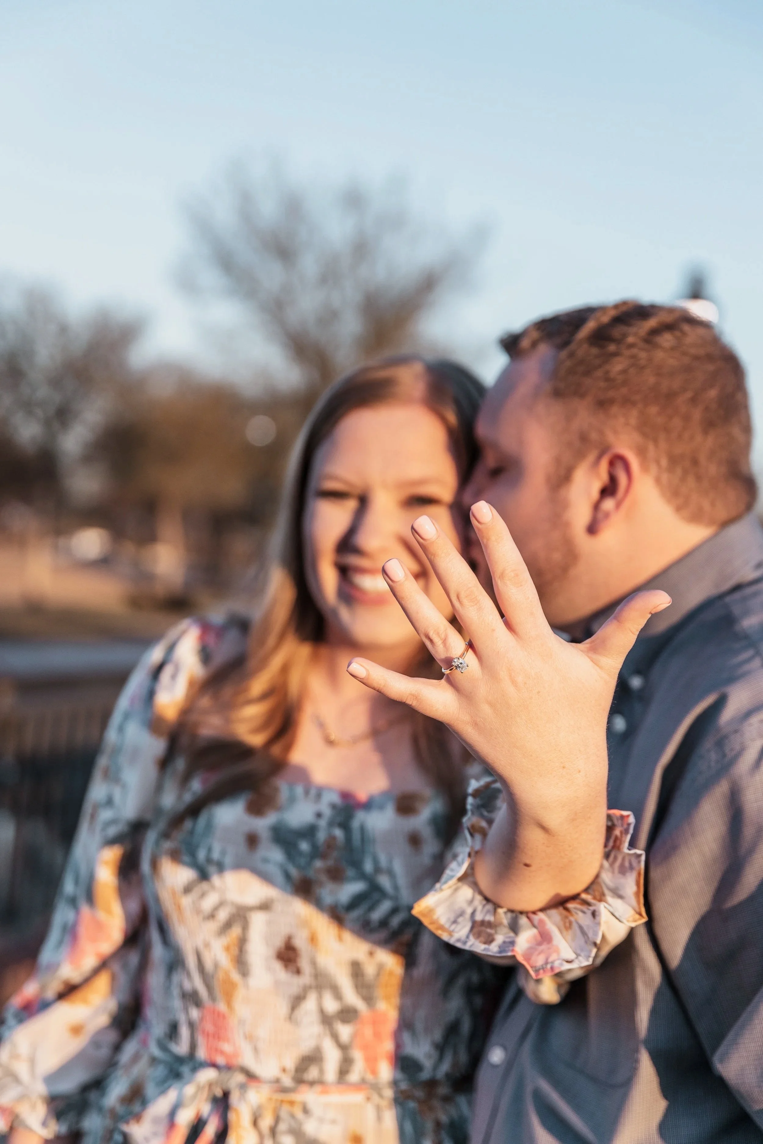A couple sharing a kiss outdoors with the woman showing her engagement ring, woman with long brown hair in a floral dress, man with short brown hair in a gray suit, blurred background of trees and sky.