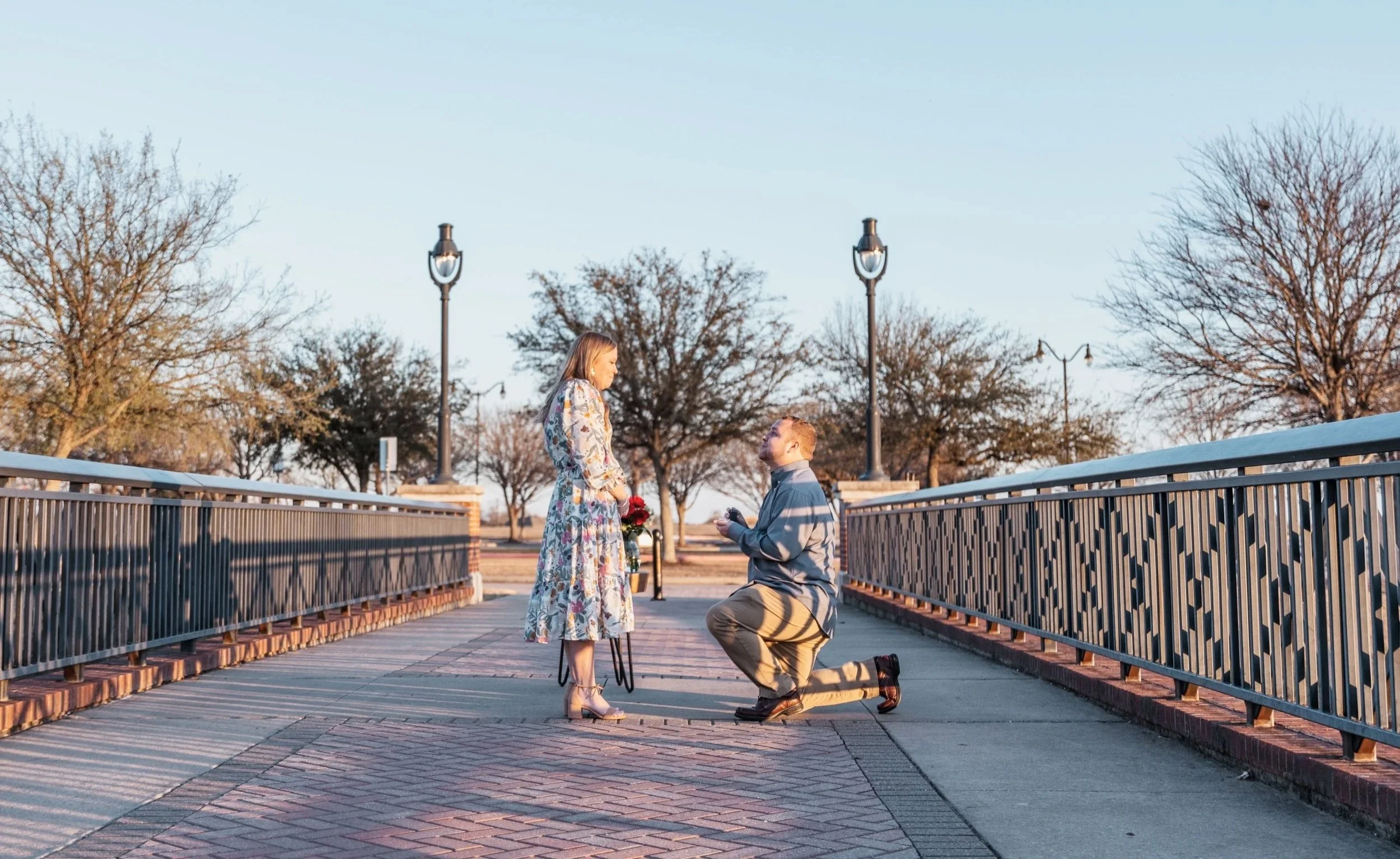 Man proposing to woman on a bridge with trees in the background during sunset.