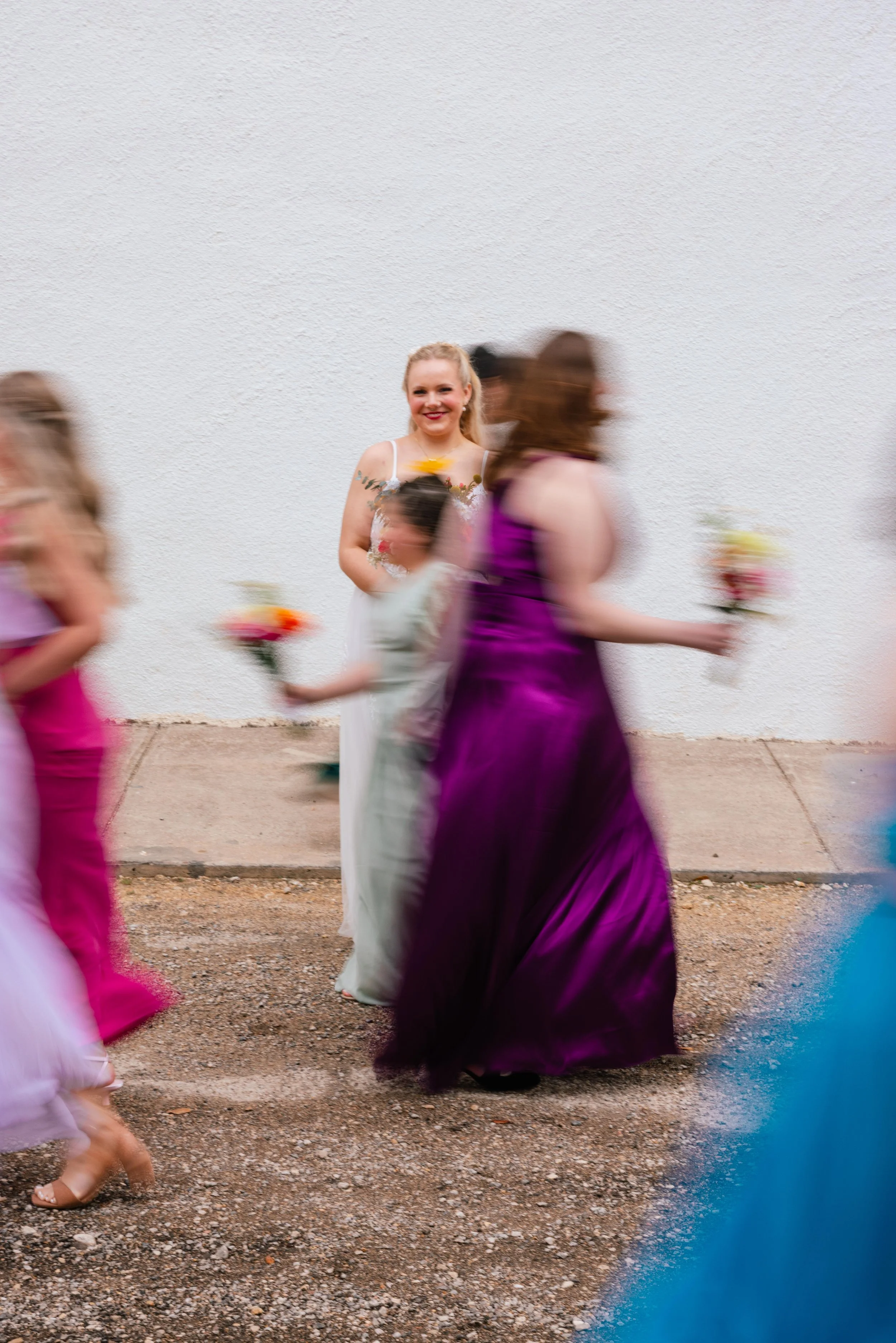 A smiling woman in a white dress stands against a white textured wall, while blurred women in purple and pink dresses walk past her holding flowers.