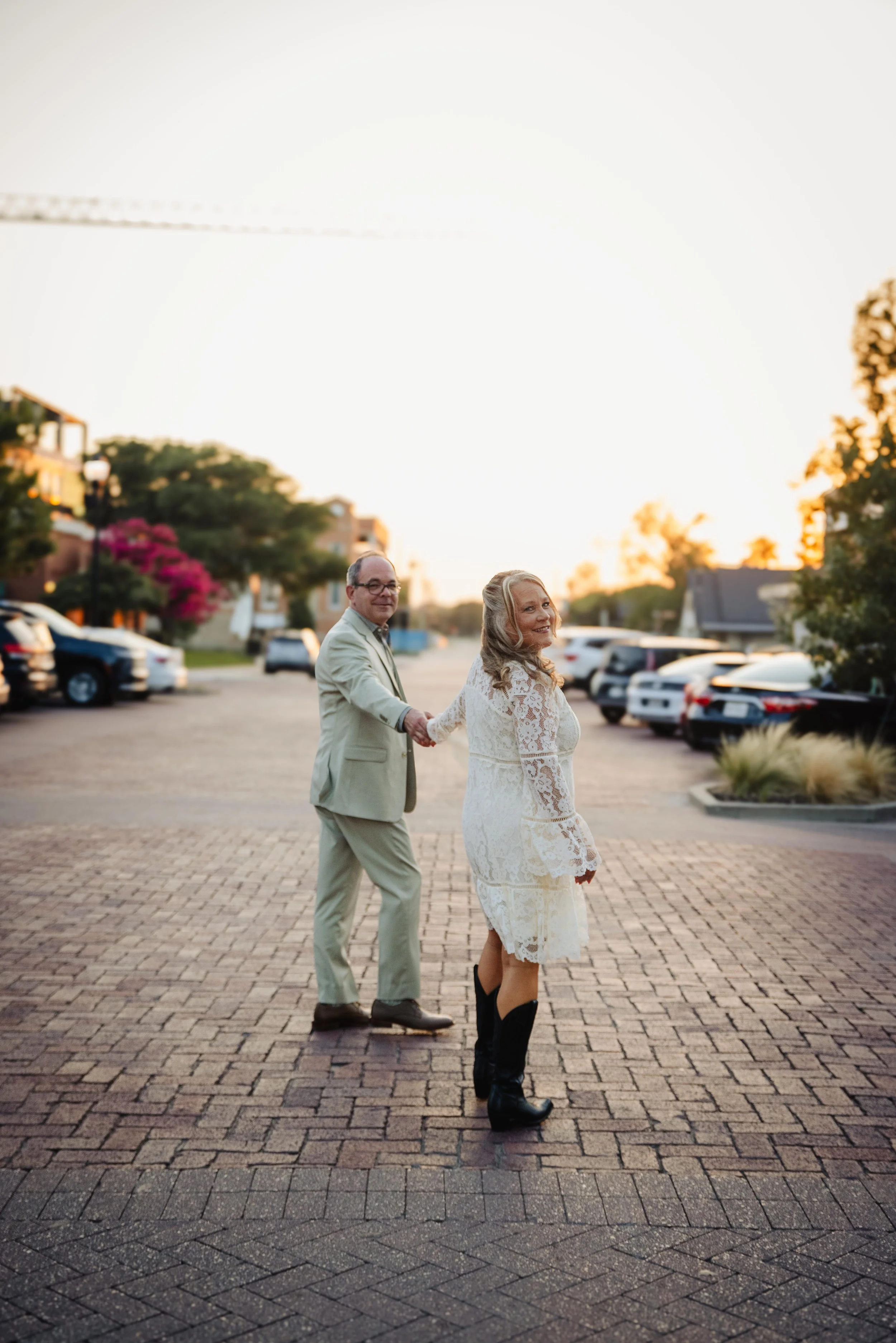 A couple holding hands and walking together outdoors on a brick sidewalk during sunset, with parked cars and trees in the background.