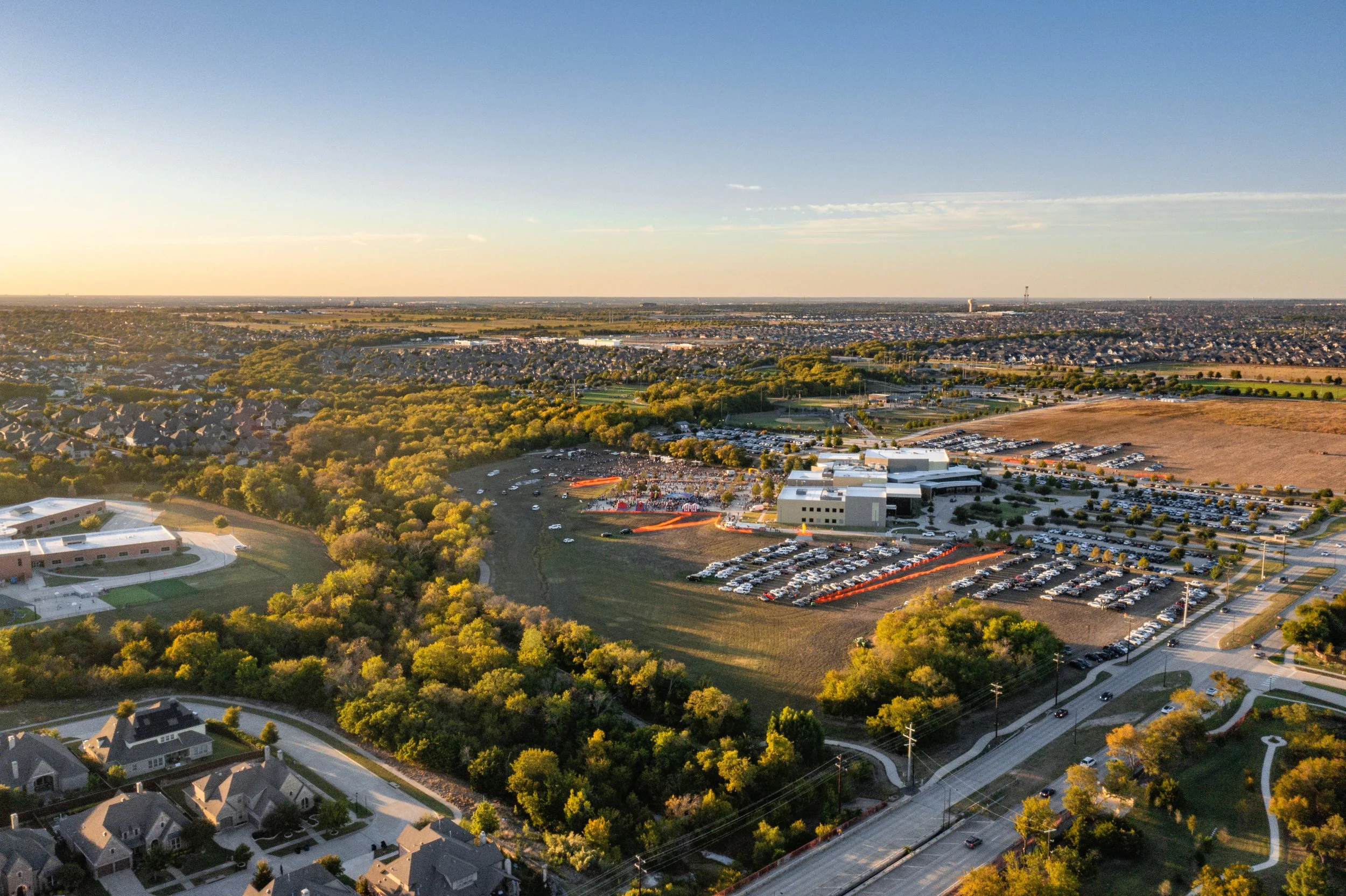 Aerial view of a suburban area with houses, trees, and parking lots, taken during sunset with a clear sky.