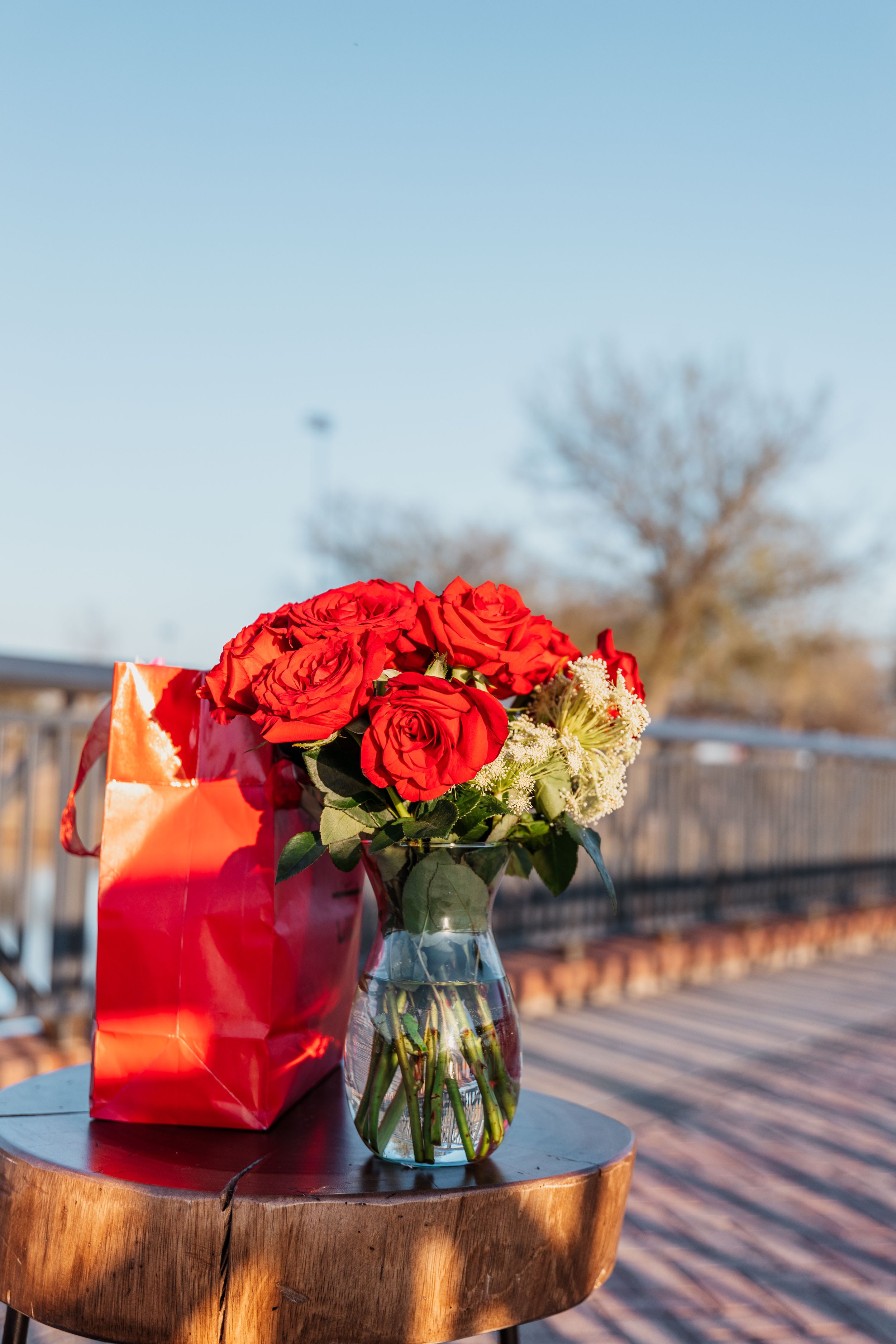 Bouquet of red roses and white flowers in a glass vase on a wooden table with a red gift bag nearby, outdoors on a clear sunny day.