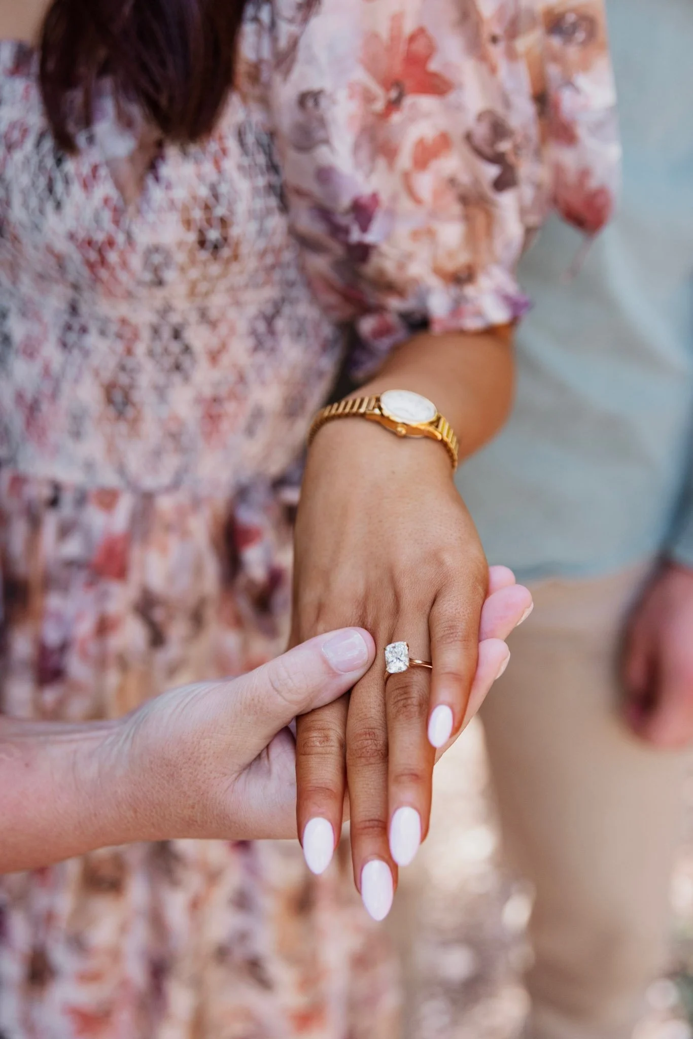 Two people holding hands, showing engagement rings, one with a gold watch, one with painted nails, and someone wearing a floral dress.