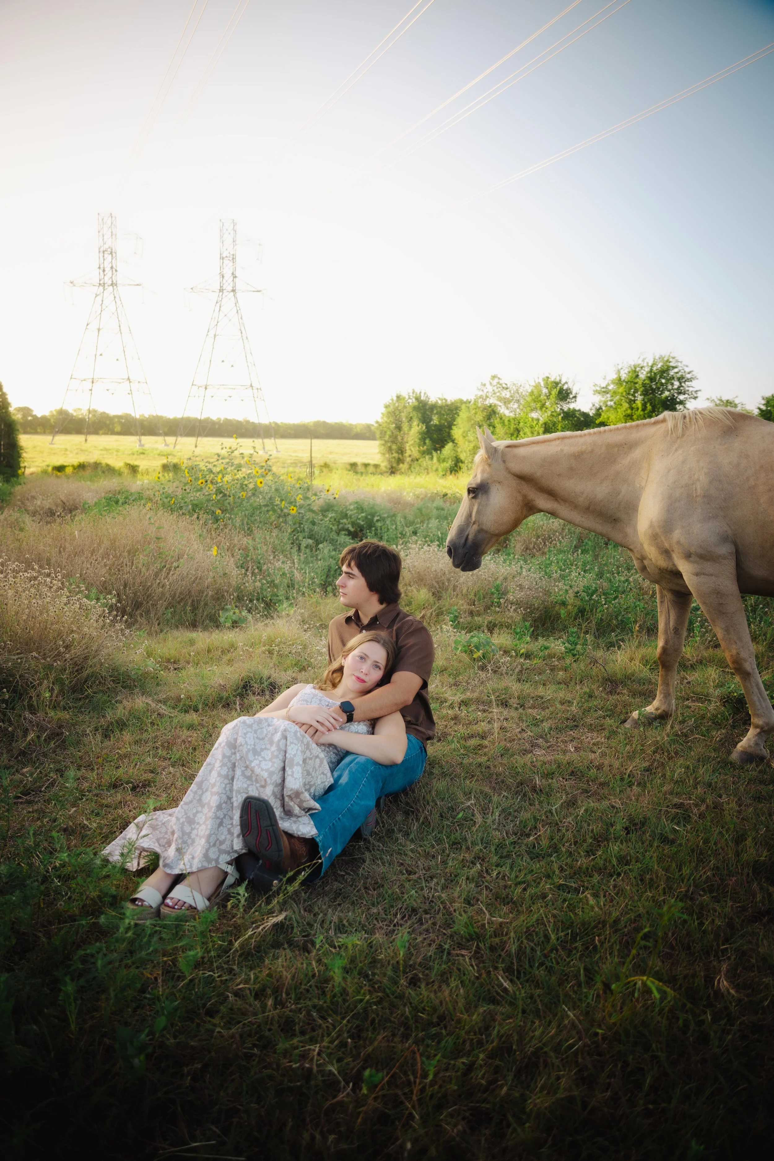 Two young people sitting on the grass in a field with a horse, trees, and power lines in the background during daylight.