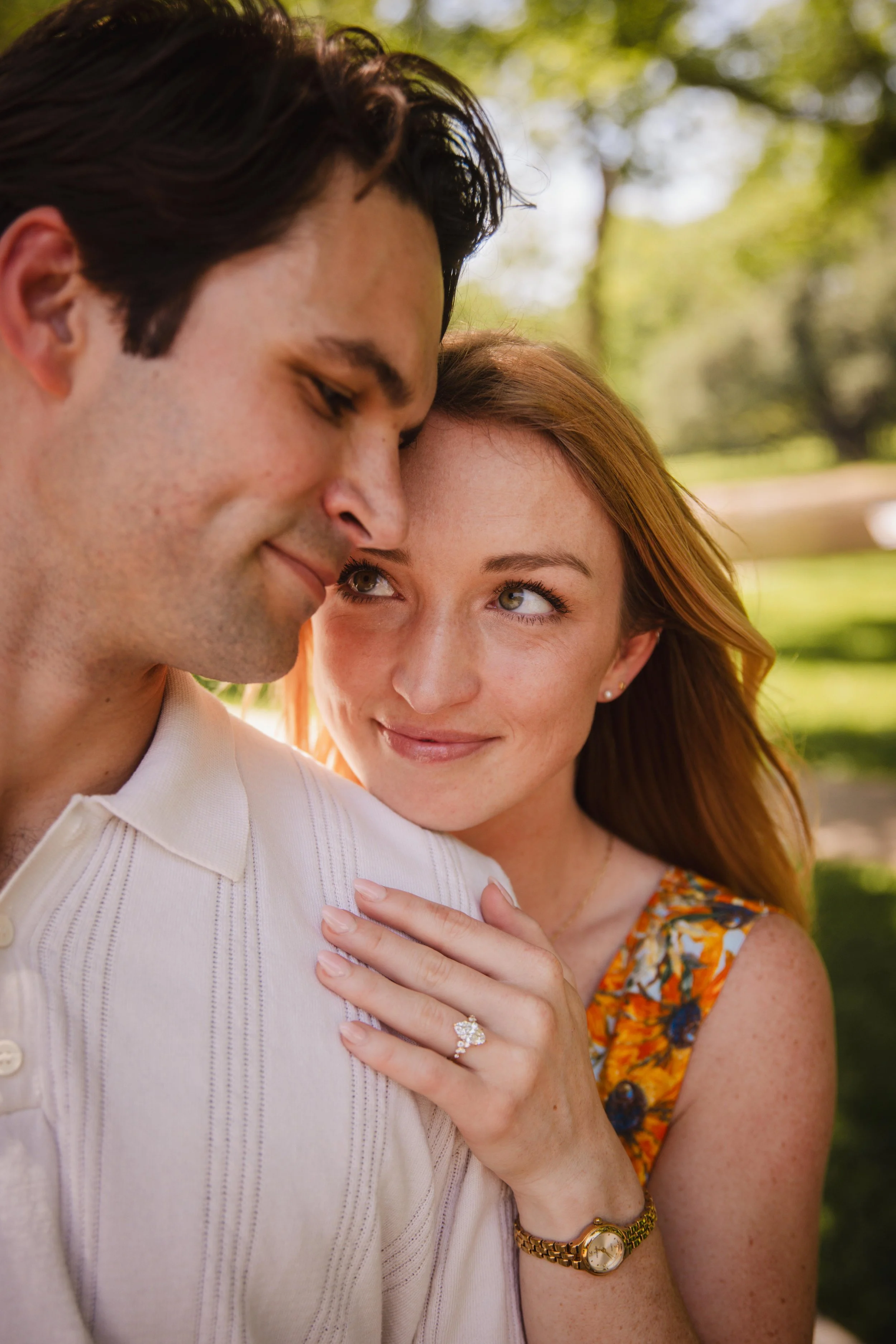 A close-up of a couple outdoors, the woman with her hand on the man's shoulder, showing an engagement ring. They are smiling and looking at each other. The background features green trees and sunlight.