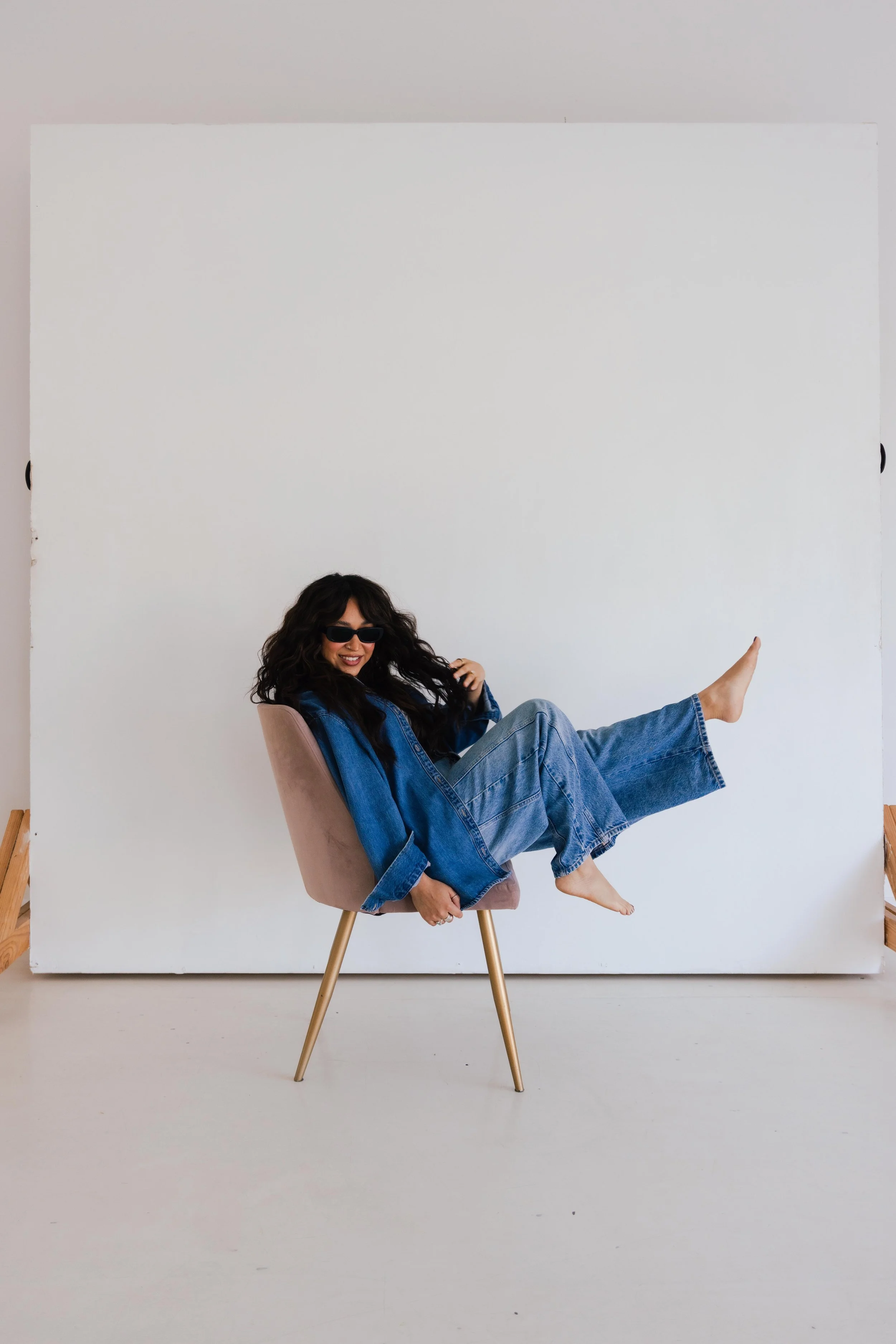 Woman with curly dark hair wearing sunglasses and a denim jacket, sitting sideways on a light pink chair, with one leg extended and one hand raised, in front of a white background.