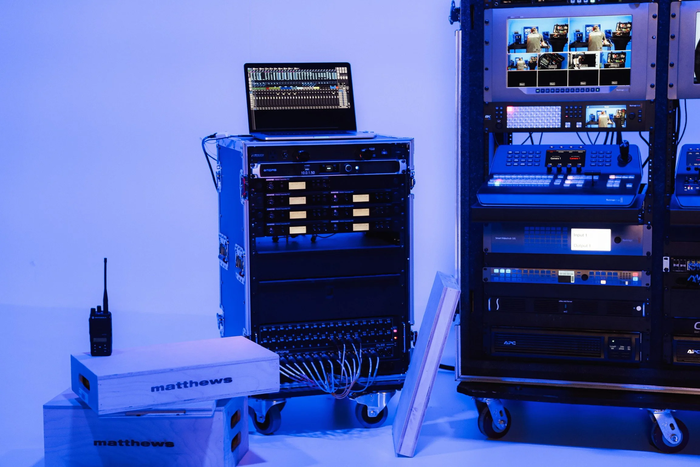 Video production equipment setup with a laptop displaying an audio mixer, a rack with audio and video hardware, and a monitor showing multiple camera feeds, all illuminated with blue lighting.