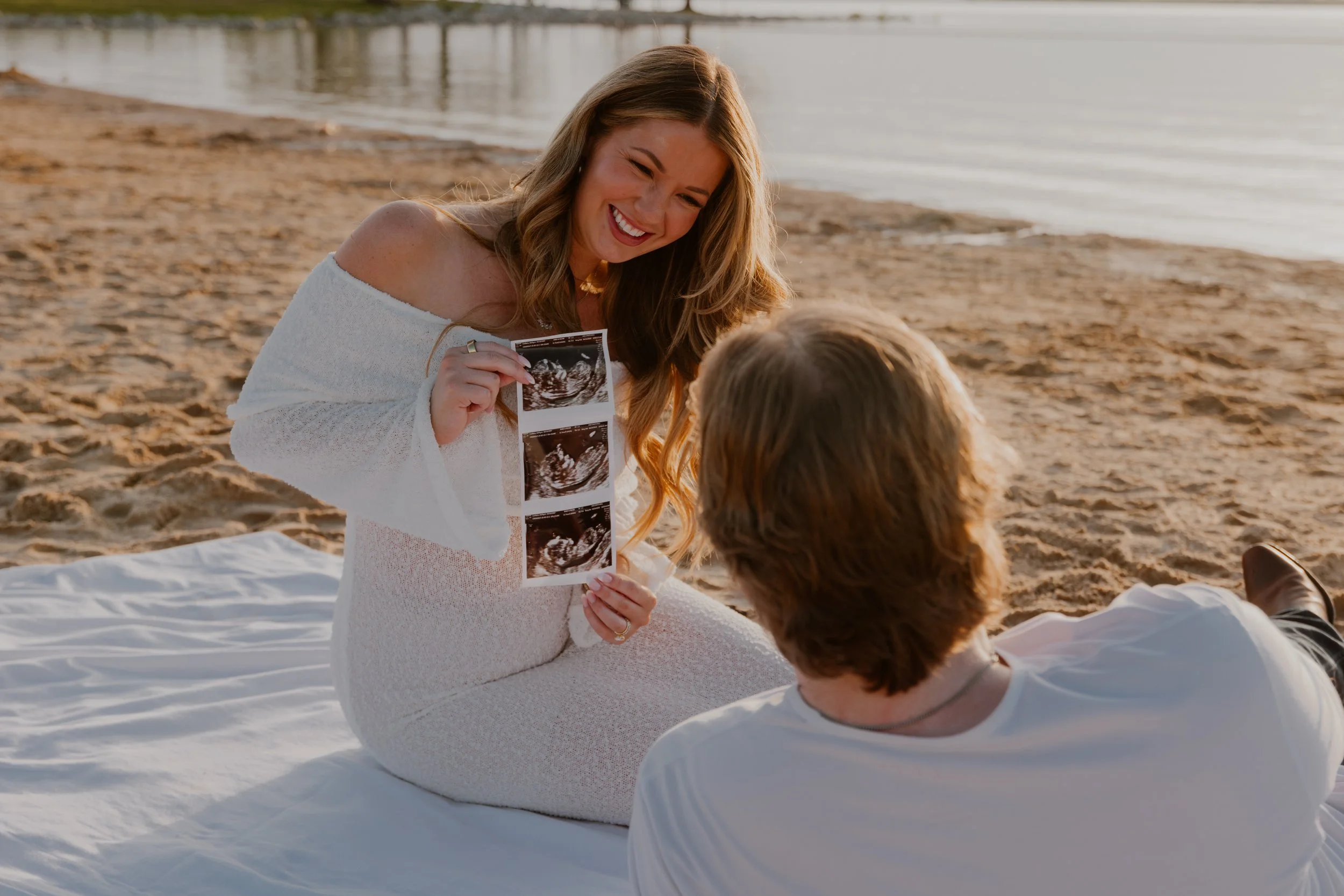 A woman with long light brown hair holding an ultrasound image, smiling at a man lying on a white blanket on the beach, with water and a pier in the background.