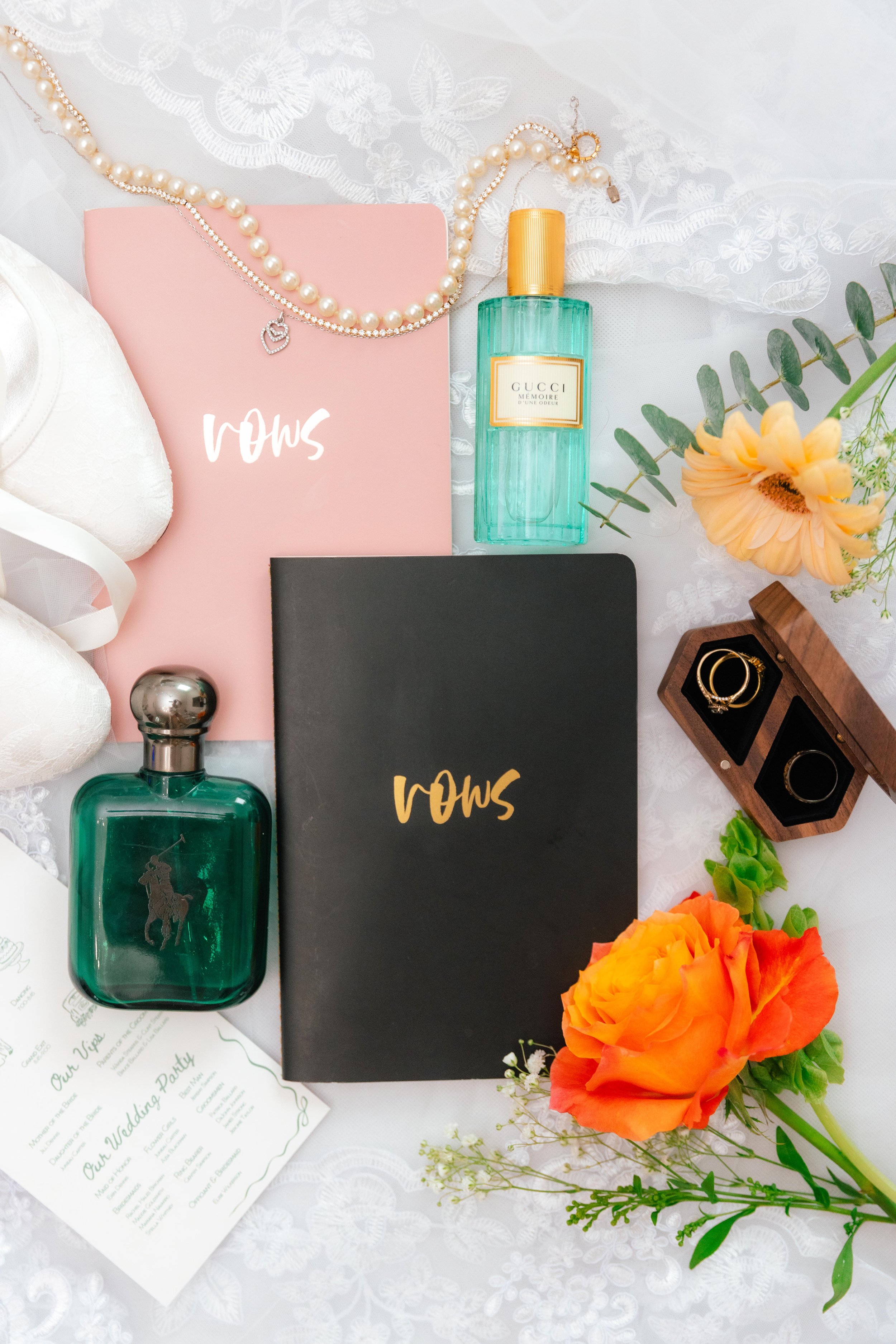 A flat lay of wedding accessories and decor on a white lace tablecloth. Includes a pink wedding planner, pearl necklace, silver necklace, a bottle of Gucci perfume, two bottles of perfume, a black notebook, a jewelry box with gold rings, a large orange flower, and smaller yellow and orange flowers.