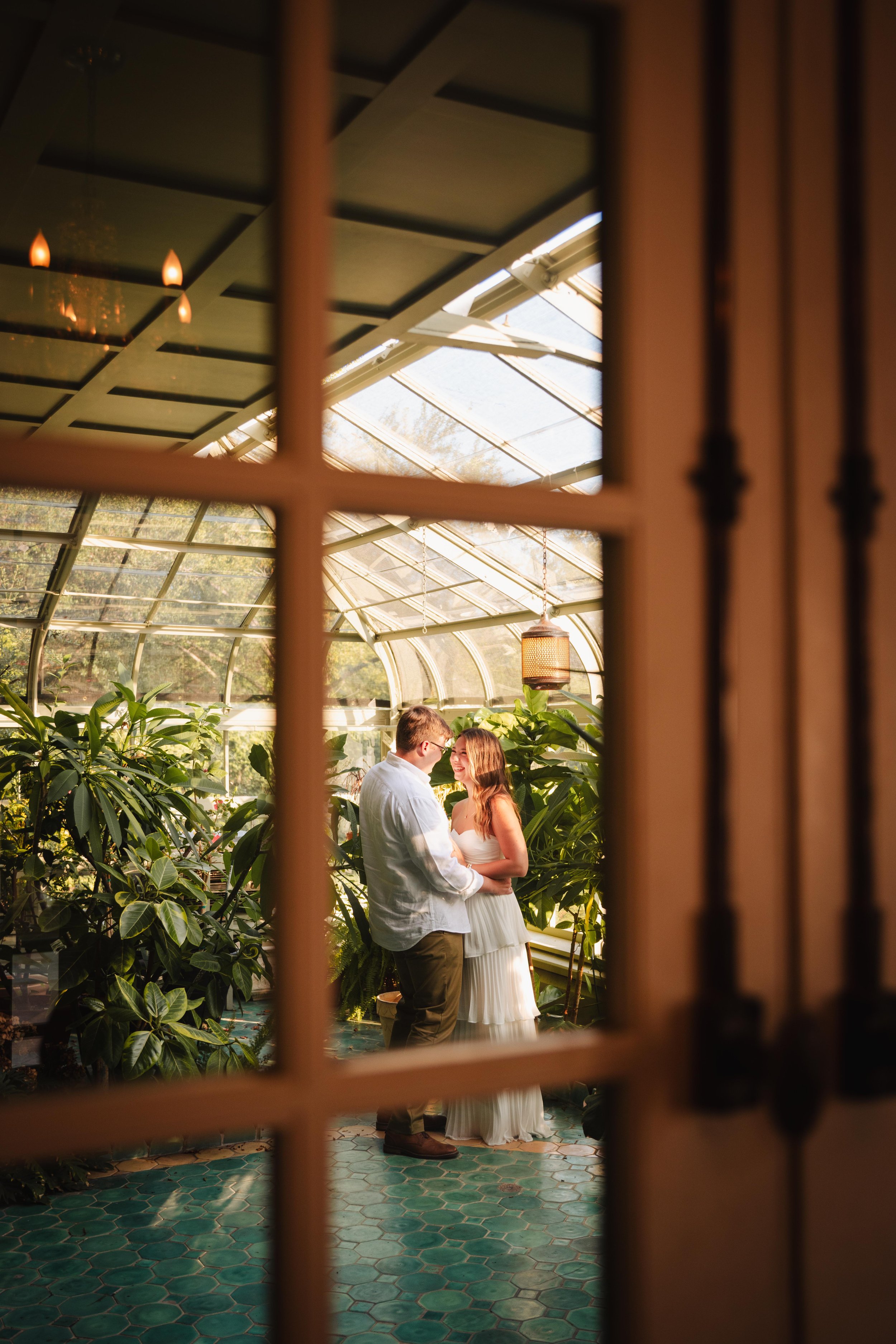 A couple in wedding attire standing inside a greenhouse or conservatory, viewed through a window with wooden framing. The man is wearing a light-colored shirt and khaki pants, and the woman is wearing a white dress. They are looking at each other and