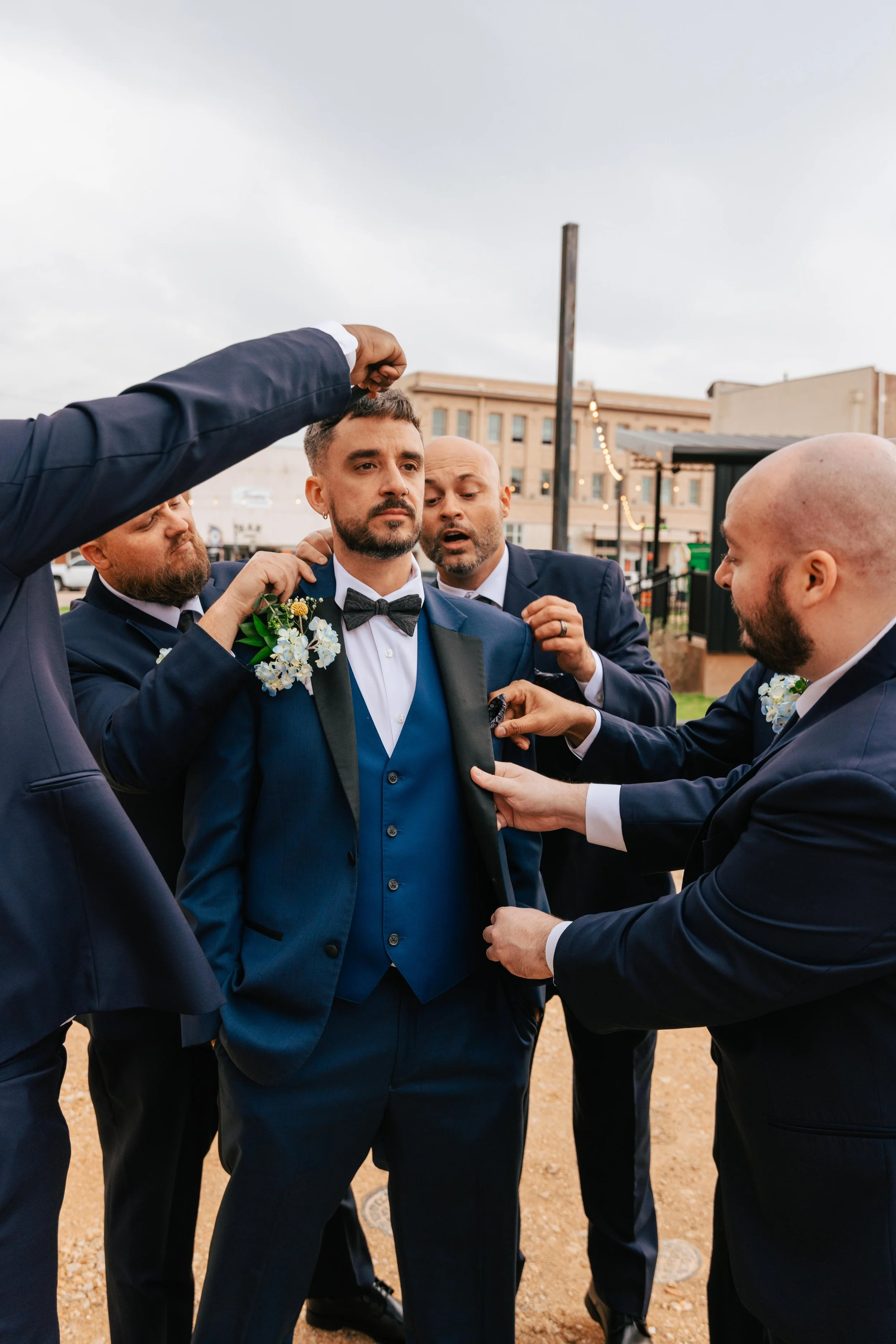 Groom in a navy blue tuxedo with a black bow tie and boutonnière, surrounded by groomsmen in suits, at an outdoor wedding ceremony, adjusting his attire.