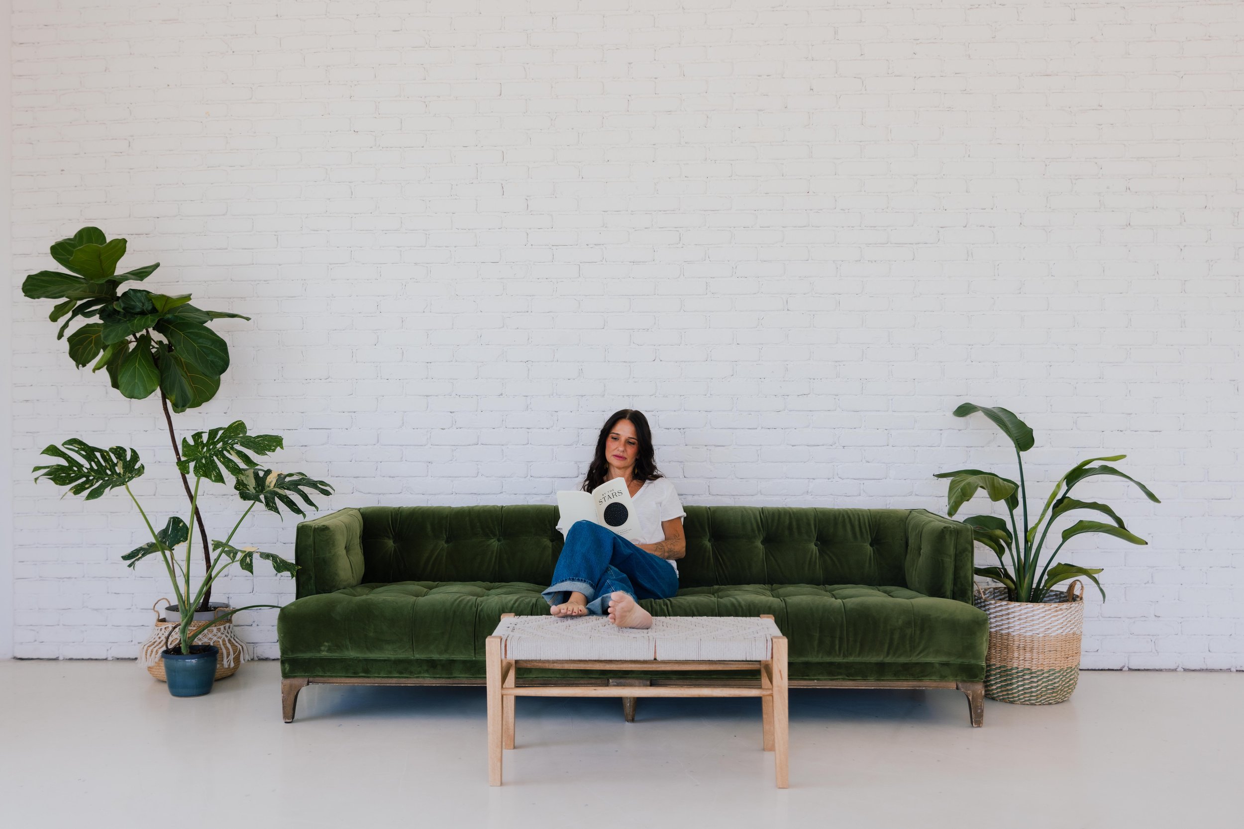 A woman sitting on a green velvet sofa reading a book in a room with a white brick wall. Several large green houseplants are placed around her.
