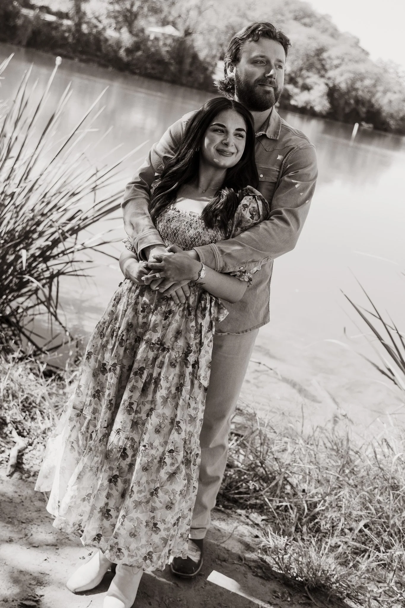 A black and white photo of a young couple standing near a body of water, the woman against the man’s chest. They are outdoors on a sandy or grassy area, with reeds and trees in the background, smiling gently.