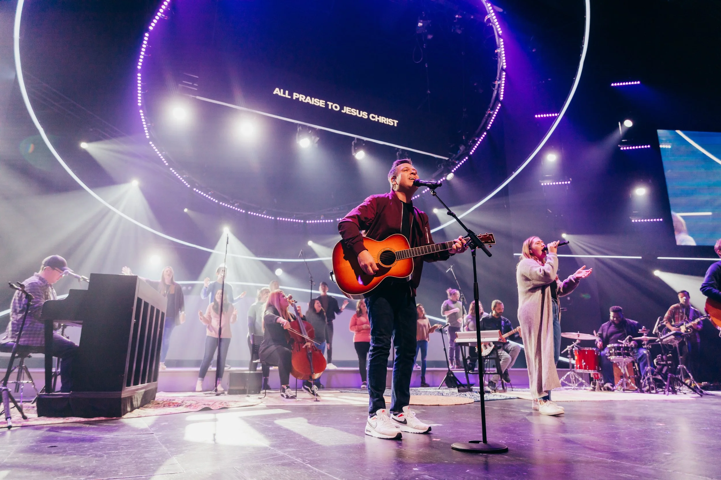 A musician playing an acoustic guitar and singing into a microphone on stage during a worship performance. A large screen above displays the words "All praise to Jesus Christ". The stage is filled with other musicians and singers, with vibrant lighti