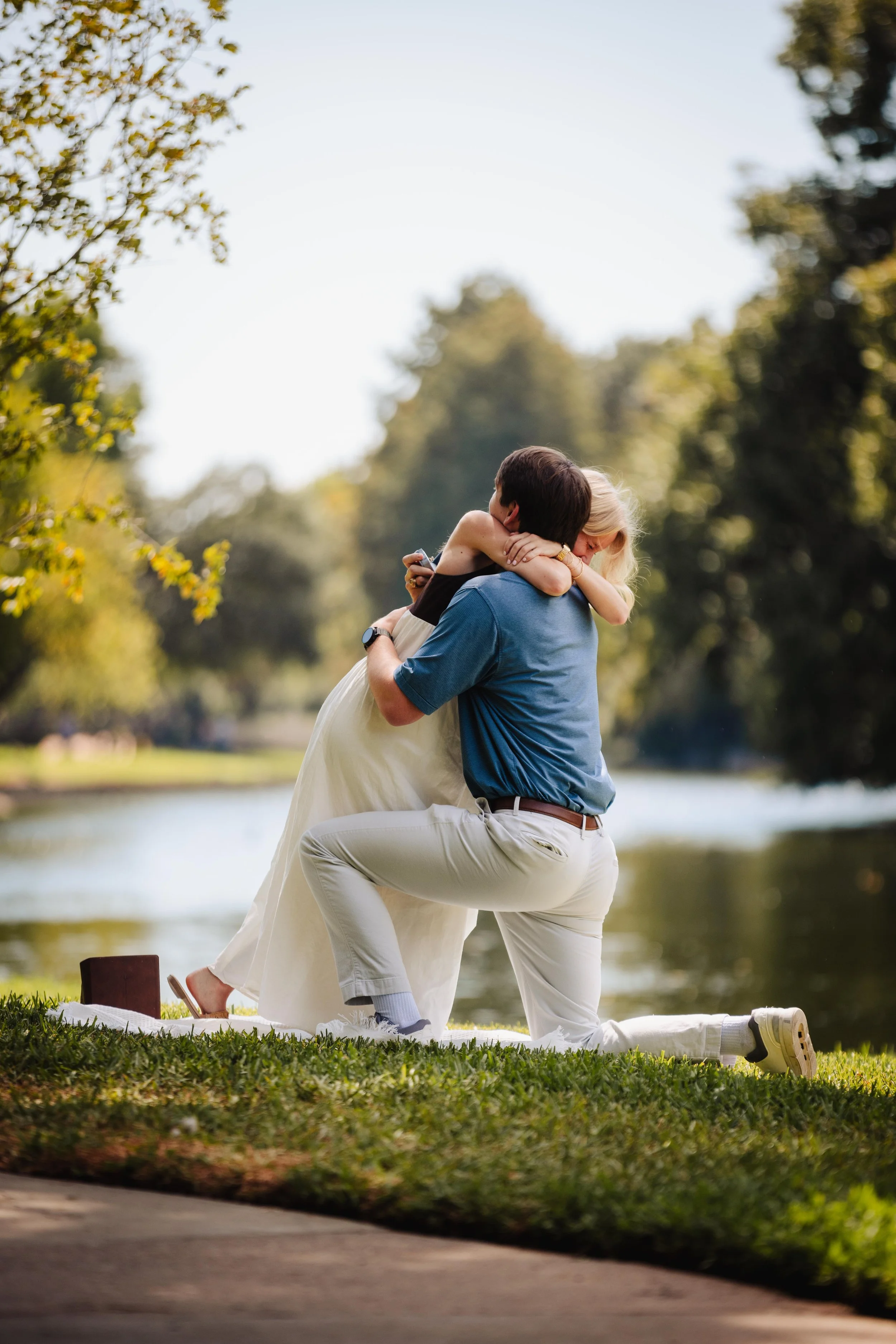 A man kneeling on grass by a river, proposing marriage to a woman who is embracing him outdoors with trees and water in the background.