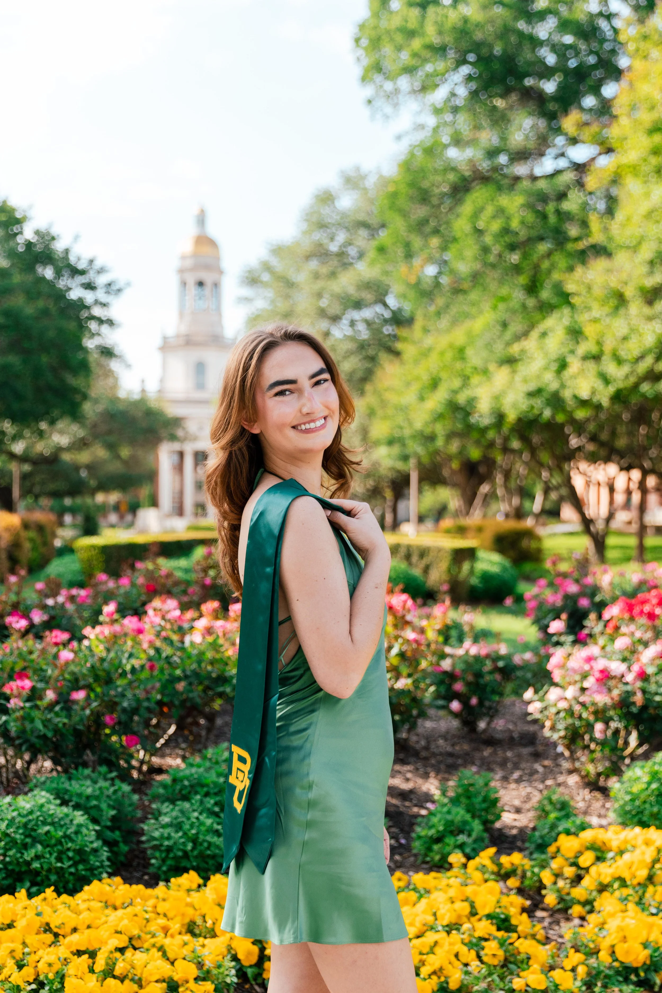 A young woman in a green dress smiling outdoors in a garden with colorful flowers and greenery, with a large building featuring a golden dome in the background.