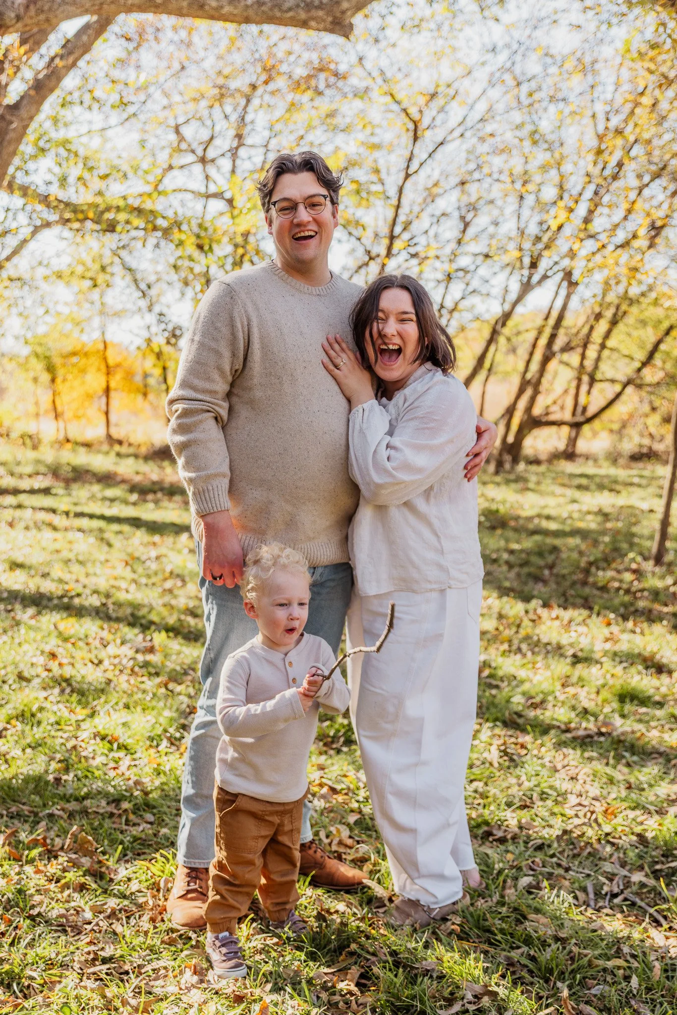 A happy family of four, including two children, enjoying time outdoors in a park during autumn. The scene captures a man and a woman smiling, with the children standing in front of them. The background features trees with yellow and orange leaves, and the ground is covered with fallen leaves.