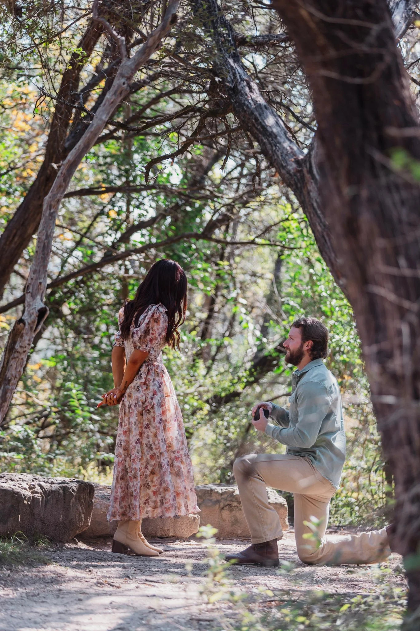 A man on one knee proposing to a woman in a forest setting, with the woman looking down at the man. The woman is wearing a floral dress and beige boots, and the man is dressed in a light blue shirt and tan pants, holding a small box. There are trees 