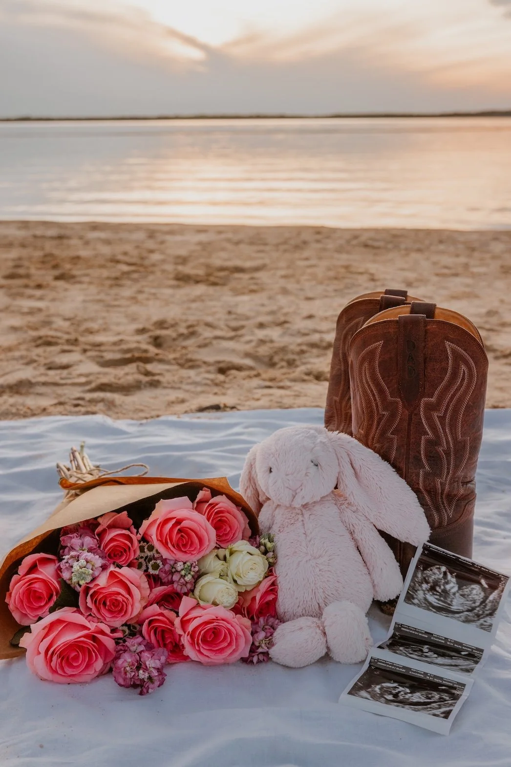 A bouquet of pink and white roses, a plush bunny toy, a pair of brown cowboy boots, ultrasound images, and a rolled blanket on a white cloth on a sandy beach during sunset.
