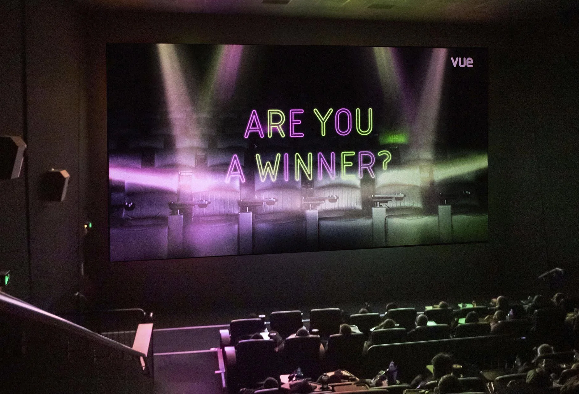 The image shows an interior of a theater or conference hall with a large screen displaying neon text that reads, 'ARE YOU A WINNER?'. The screen has a dark background with colorful lighting effects, and there are seats with people sitting in the lowe