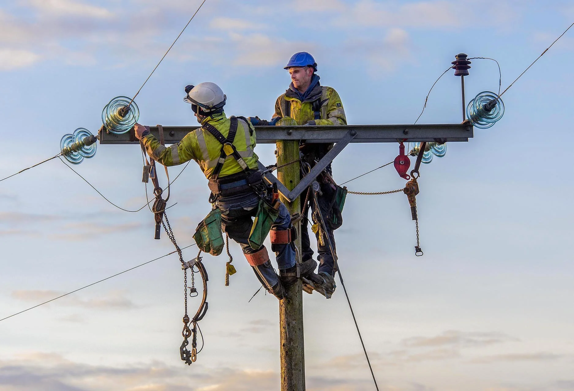 Two workers in safety gear working on power lines atop a utility pole against a cloudy sky.