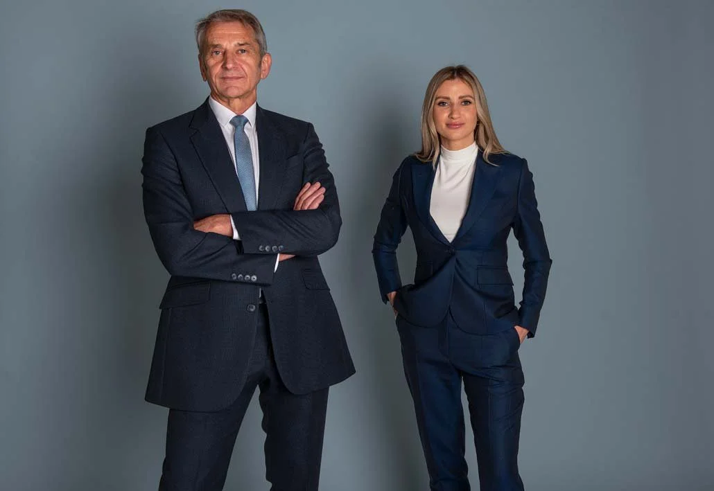 Businessman with arms crossed standing beside a businesswoman in a navy blazer, corporate duo portrait against a grey backdrop
