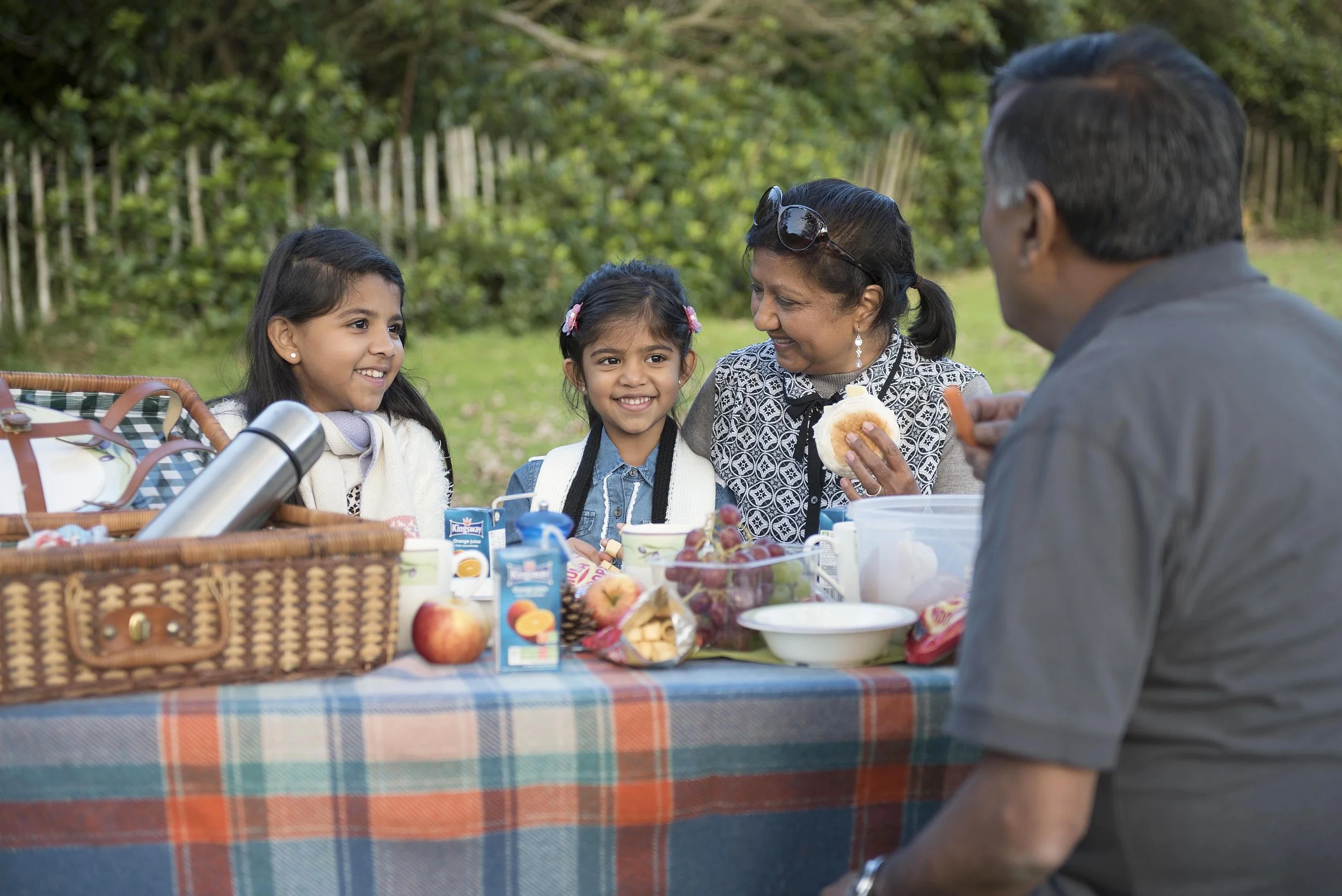 two young girls having a picnic on a table in the park with grand parents. picnic blanket and basket are clearly shown. 
