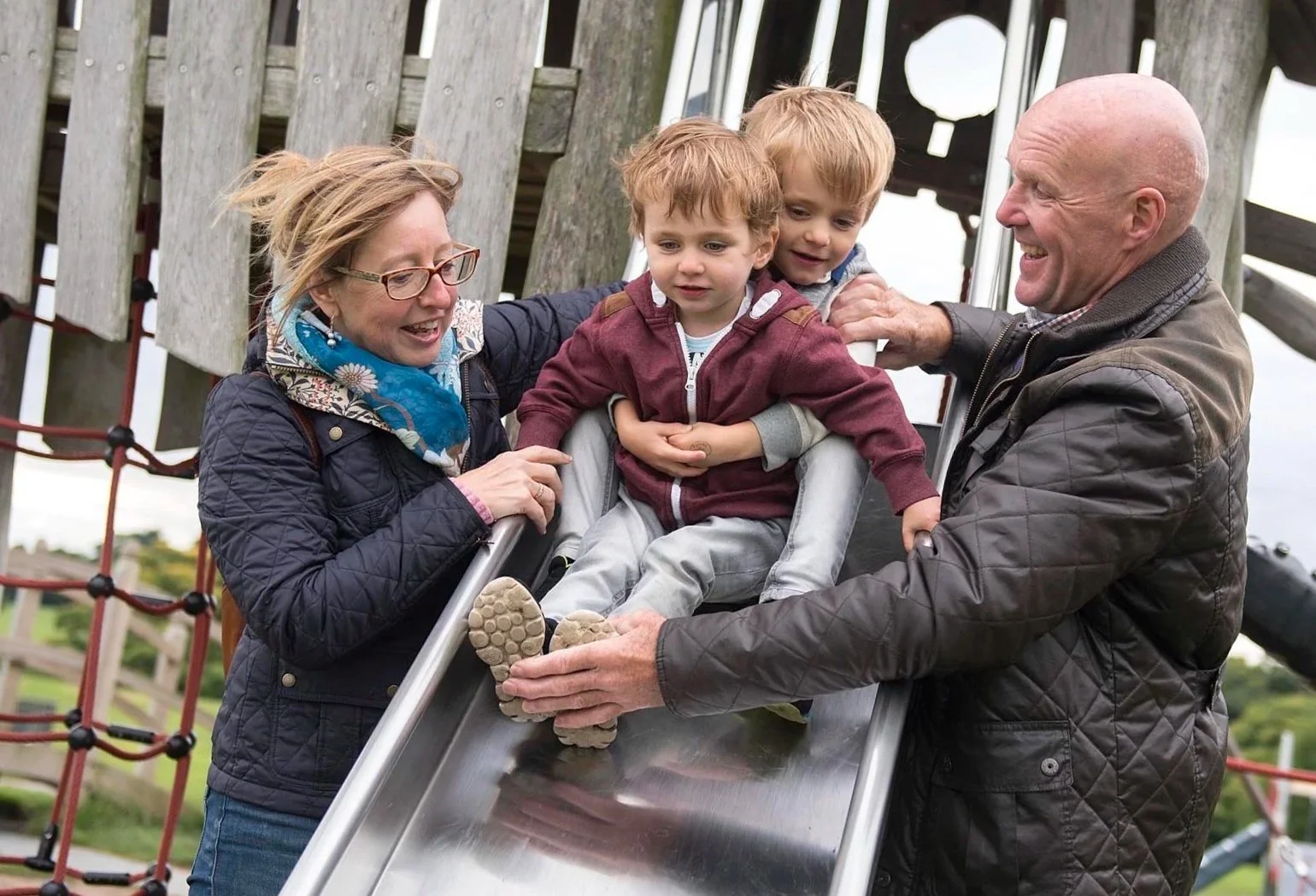 A family of four enjoying time on a playground slide outdoors. The mother and father are amid the slide, helping two young boys, one sitting on the slide in a red hoodie and gray pants, and the other slightly behind him. The family is smiling and app