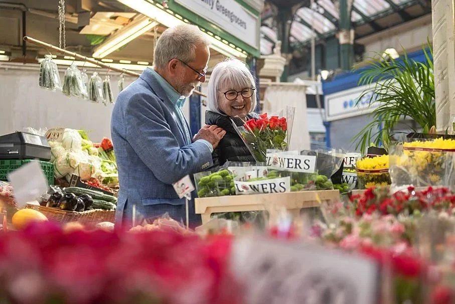 An elderly couple shopping for flowers in an indoor market, woman holding a bouquet of red roses, smiling at each other. Leeds market