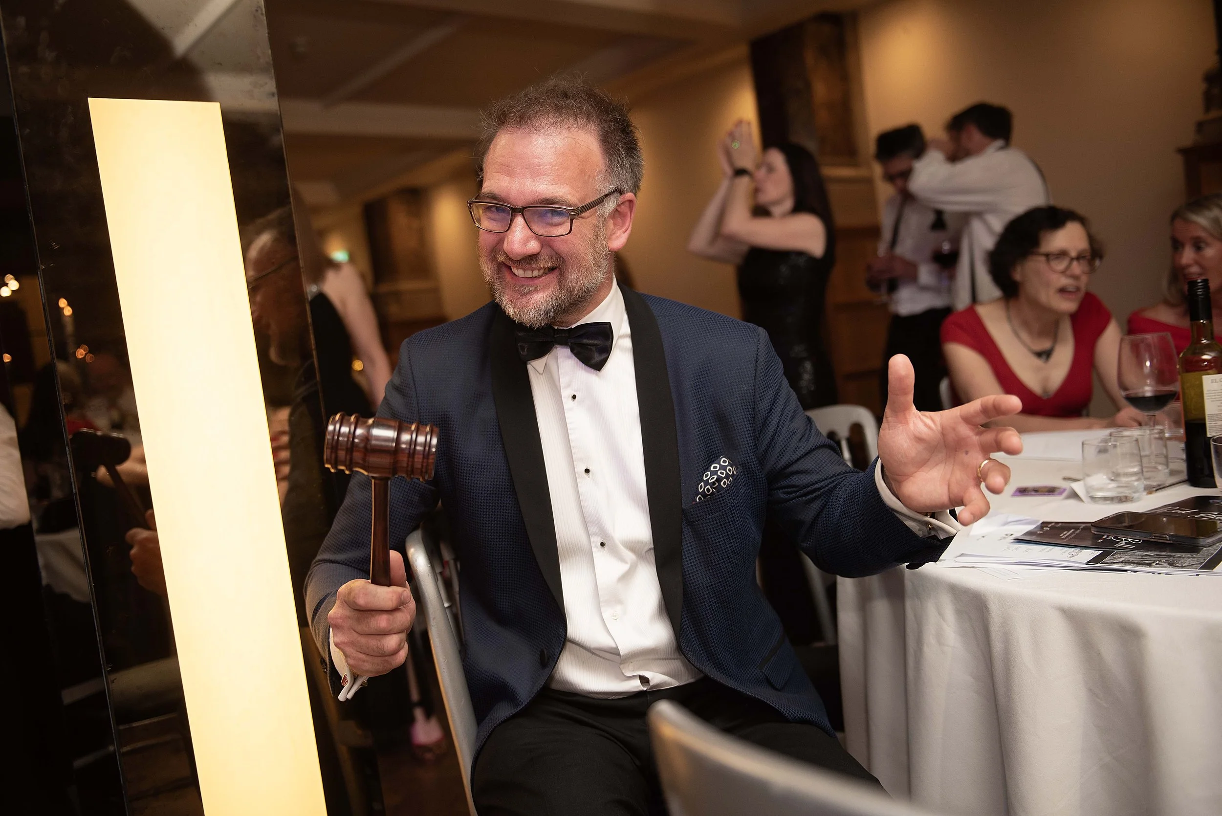 a man wearing a bow tie and dinner suit holding an auctioneers hammer and his right hand outstretched smiling. he also has a beard