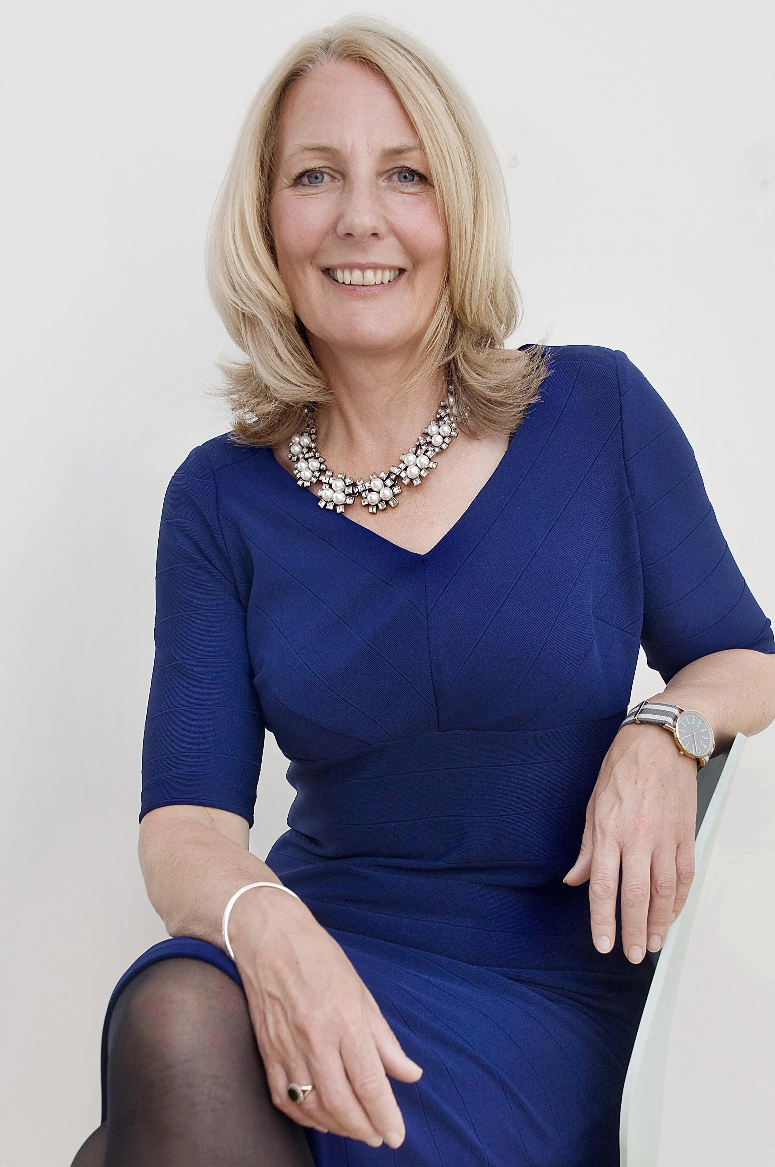 studio headshot of a woman in a blue dress sat on a chair with one arm resting on the chair. Corporate headshot photography session in Leeds studio.
