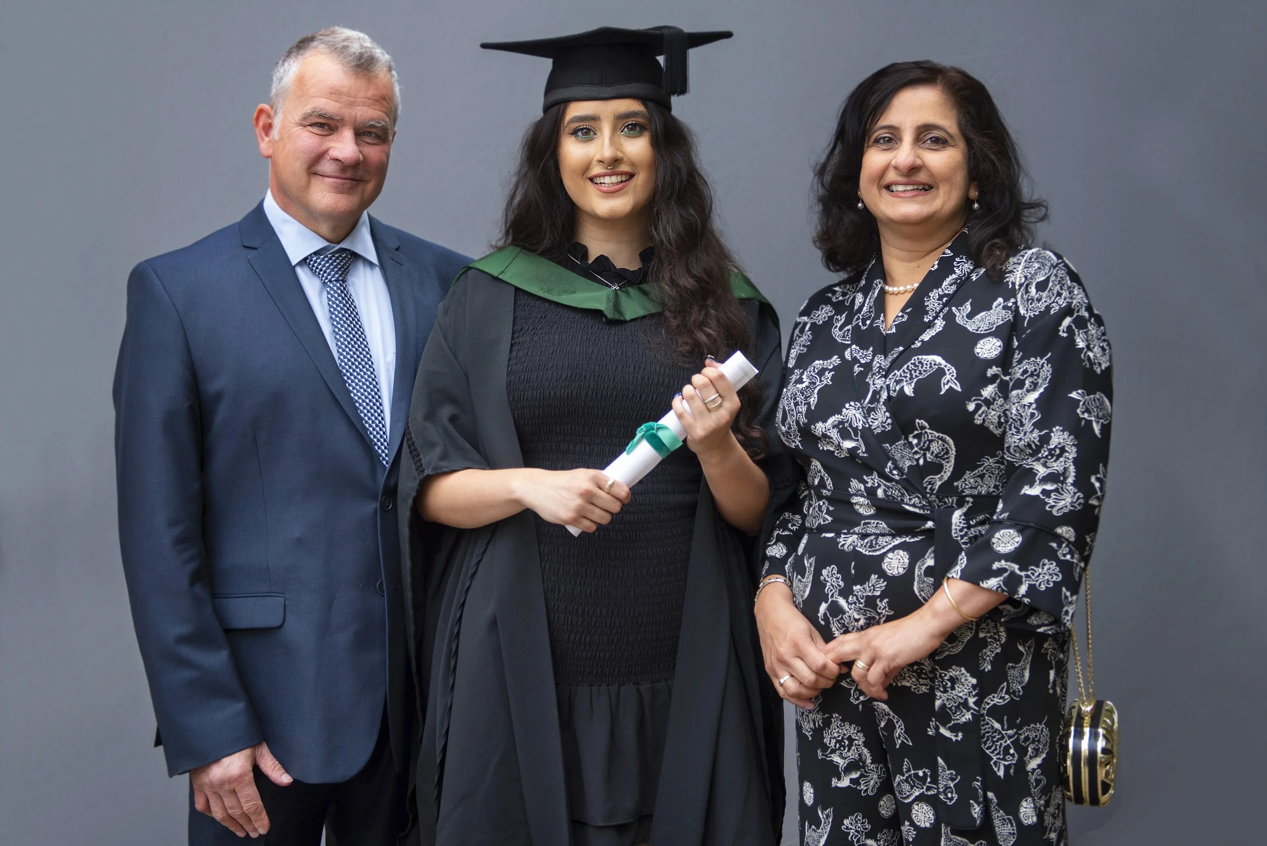 young woman and her parents in smart outfits posing with mortar board, certificate and graduation gown on grey background. Studio style lighting