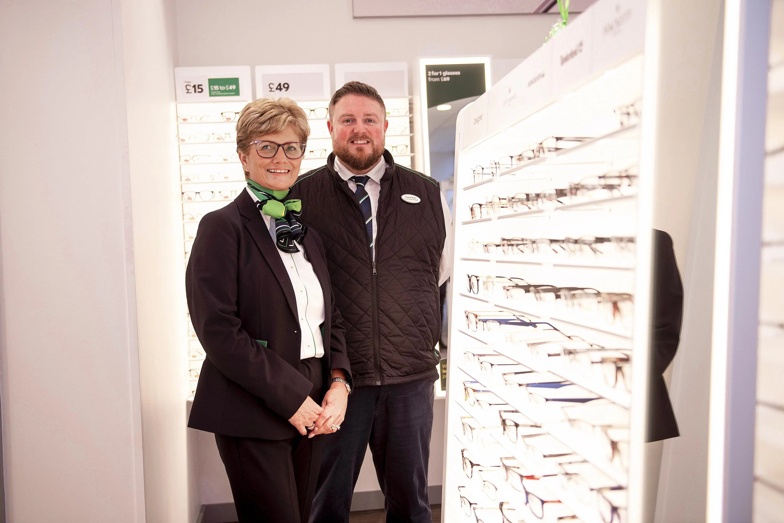 Two smiling staff members, a woman wearing glasses and a man, standing inside an eyewear store with shelves of glasses behind them.