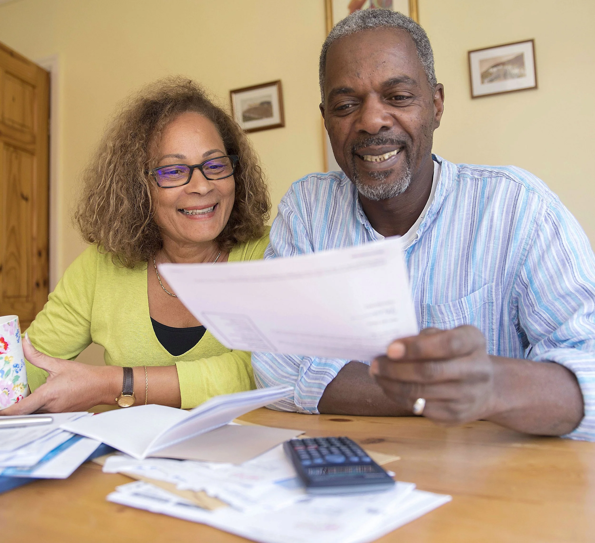 Two smiling adults, a woman with curly hair and glasses and a man with gray hair, are reading and discussing papers at a table cluttered with documents, a calculator, and a mug, in a room with light-colored walls and framed pictures.