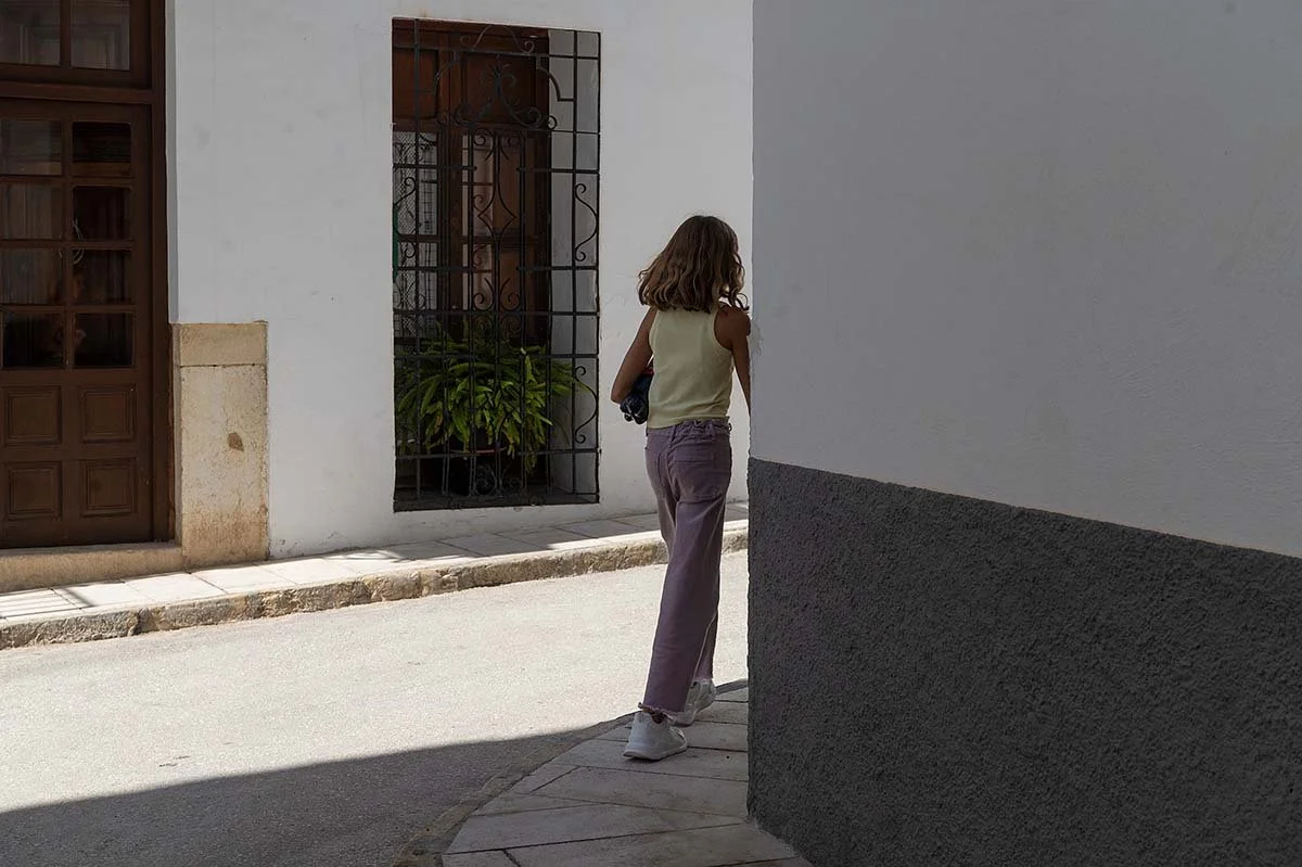 A young girl with curly hair wearing a sleeveless top, loose pants, and sneakers walking down a narrow street past a white wall, with a potted plant visible through a window with decorative bars.