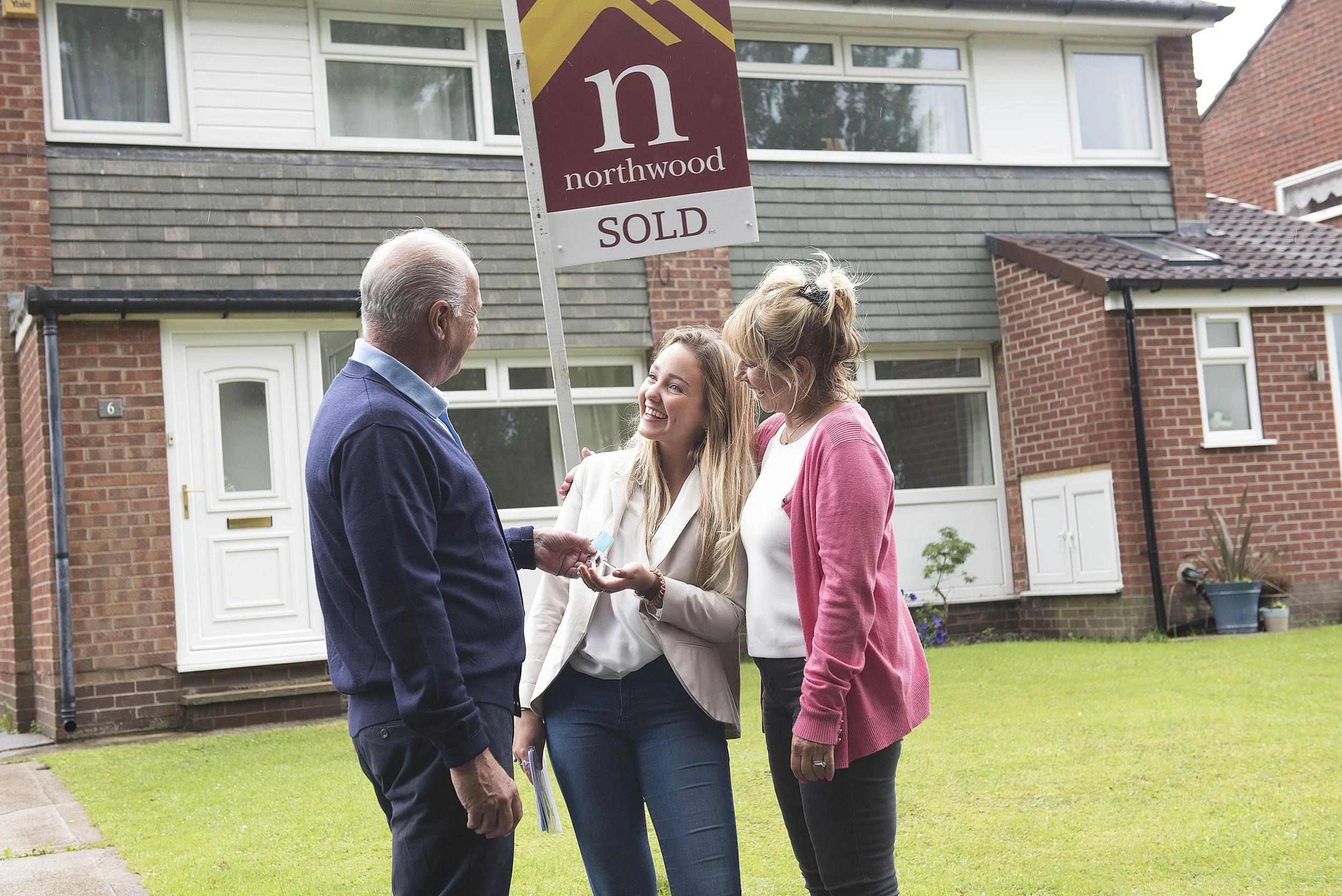 a young woman is being given the keys to her new house by an older couple. a semi detached house and sold sign are in the background. studio light quality on location in Leeds