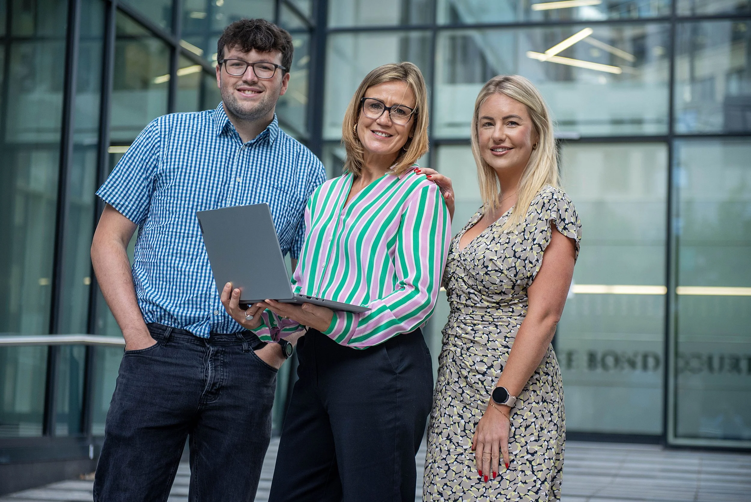 Three people standing outside a modern glass building, smiling at the camera. The man on the left wears glasses and a blue checkered shirt, holding a laptop. The woman in the middle has blonde hair, wears glasses, and a colorful striped blouse, with 