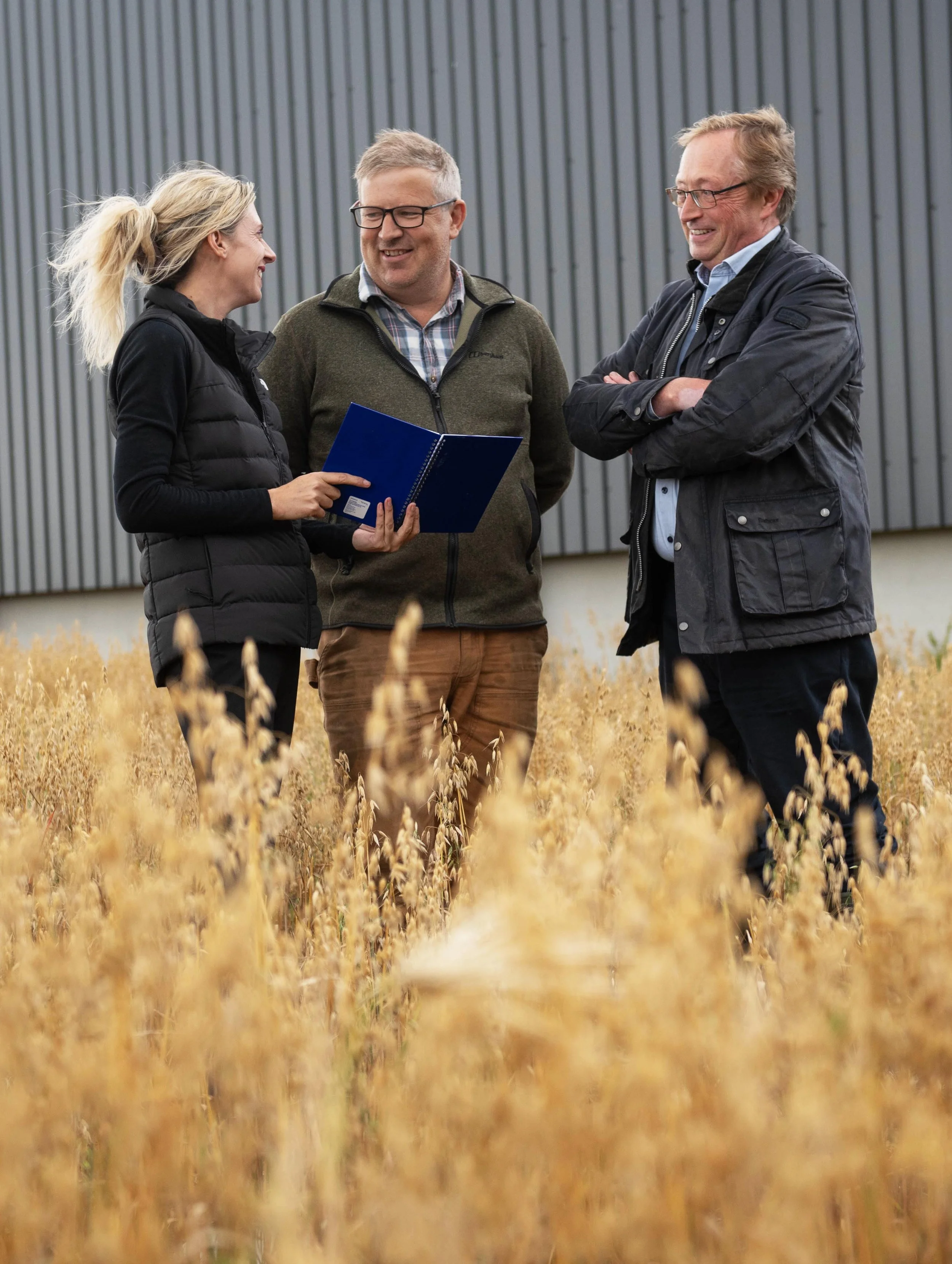 Three people standing in a wheat field, smiling and talking, with a large gray metal building in the background, two men and one woman.