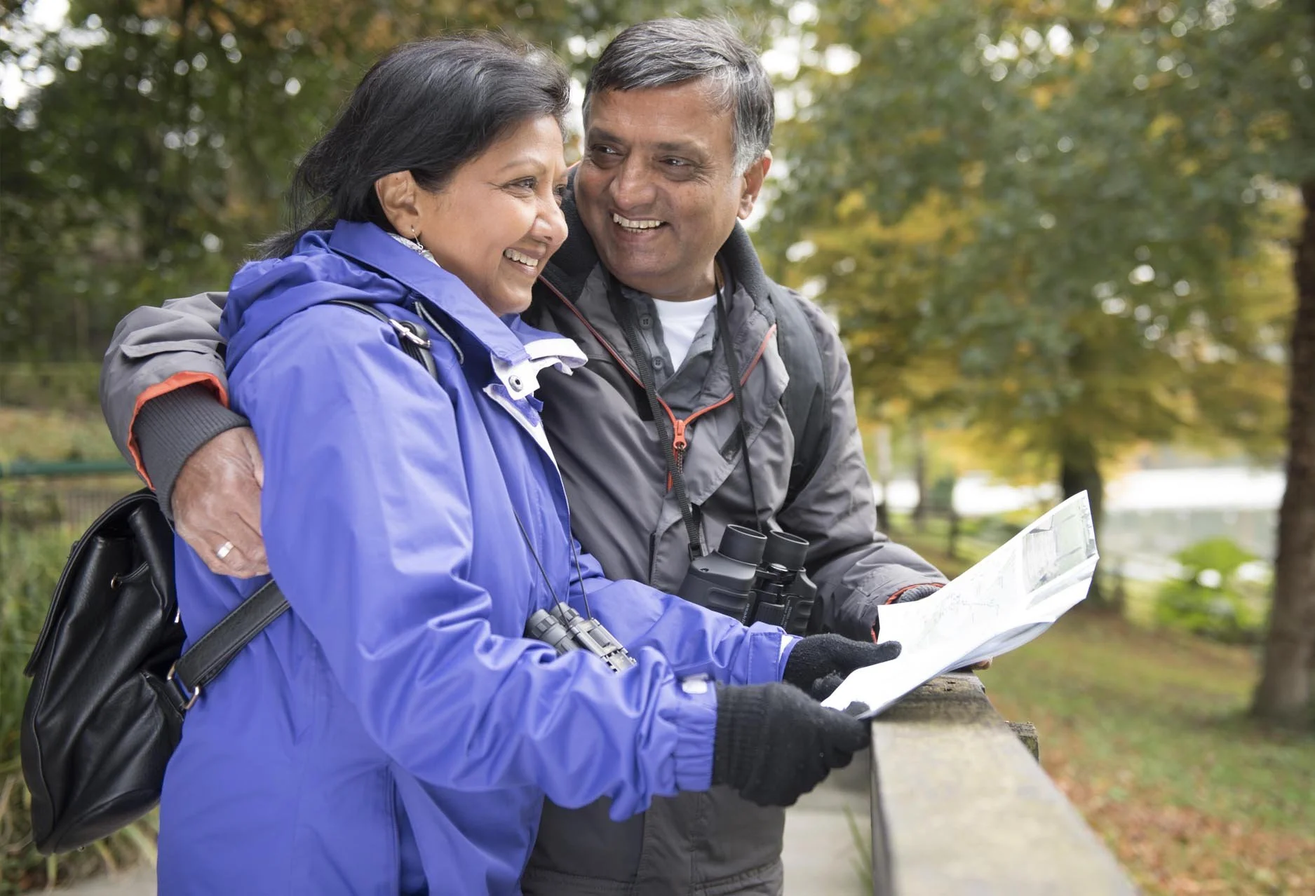 A smiling middle-aged man and woman sit on a park bench, looking at each other, with the man holding binoculars around his neck and a map in his hands, amidst a background of trees with autumn leaves.