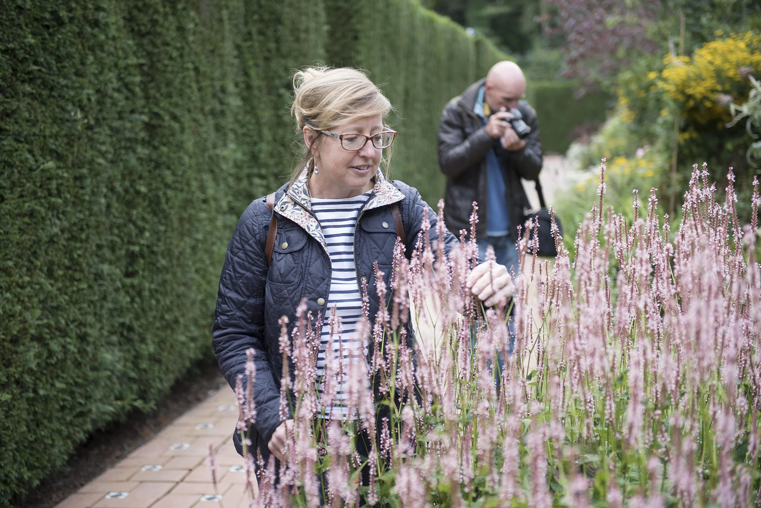 a man and woman in a public gardens. she is looking at flowers and he is taking a picture with a camera in the background