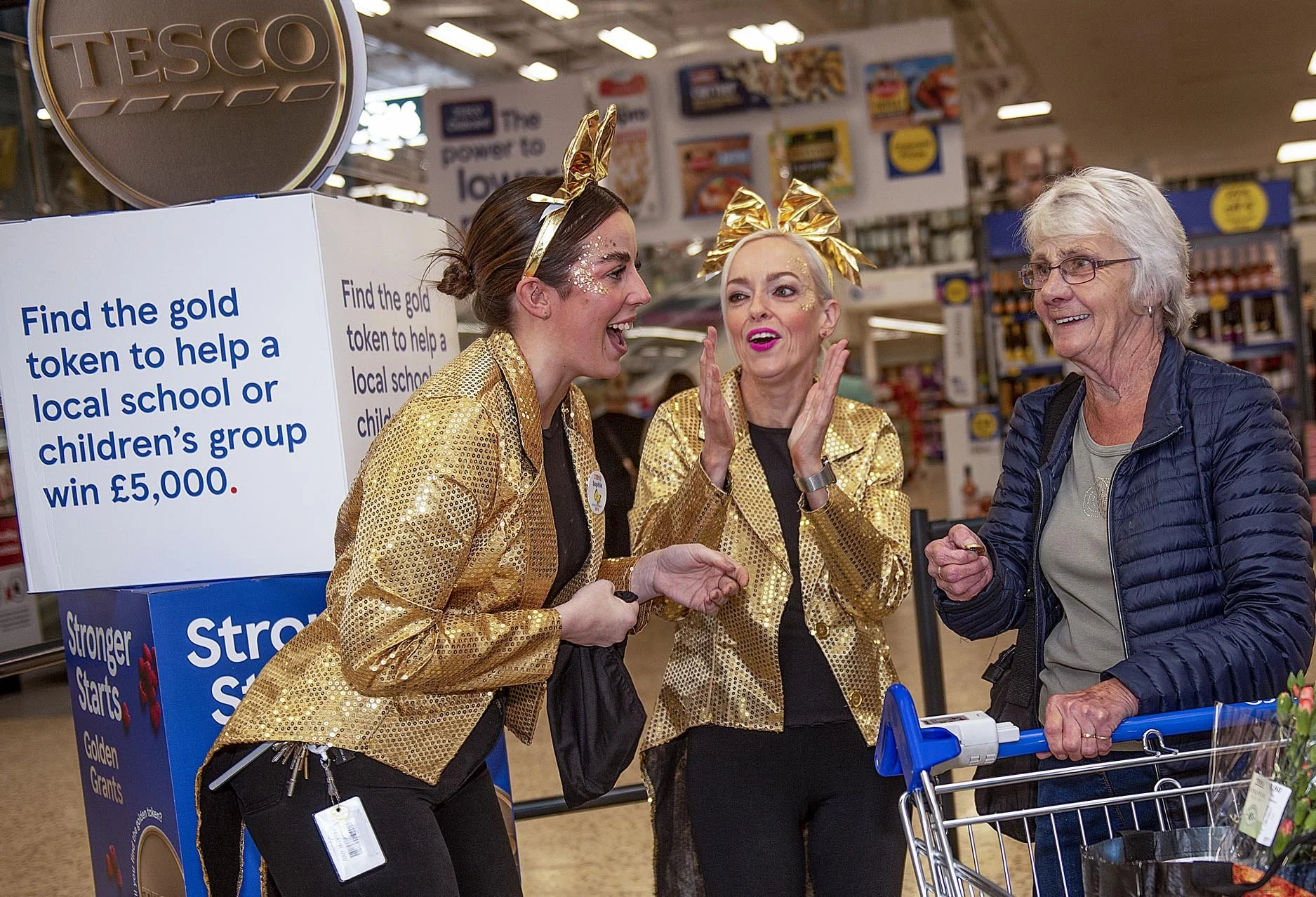 Two women dressed in gold and one woman dressed in dark clothing talking to a customer at a Tesco store. The women in gold are wearing matching gold jackets and headbands with gold bows. The customer is smiling and holding onto a shopping cart.
