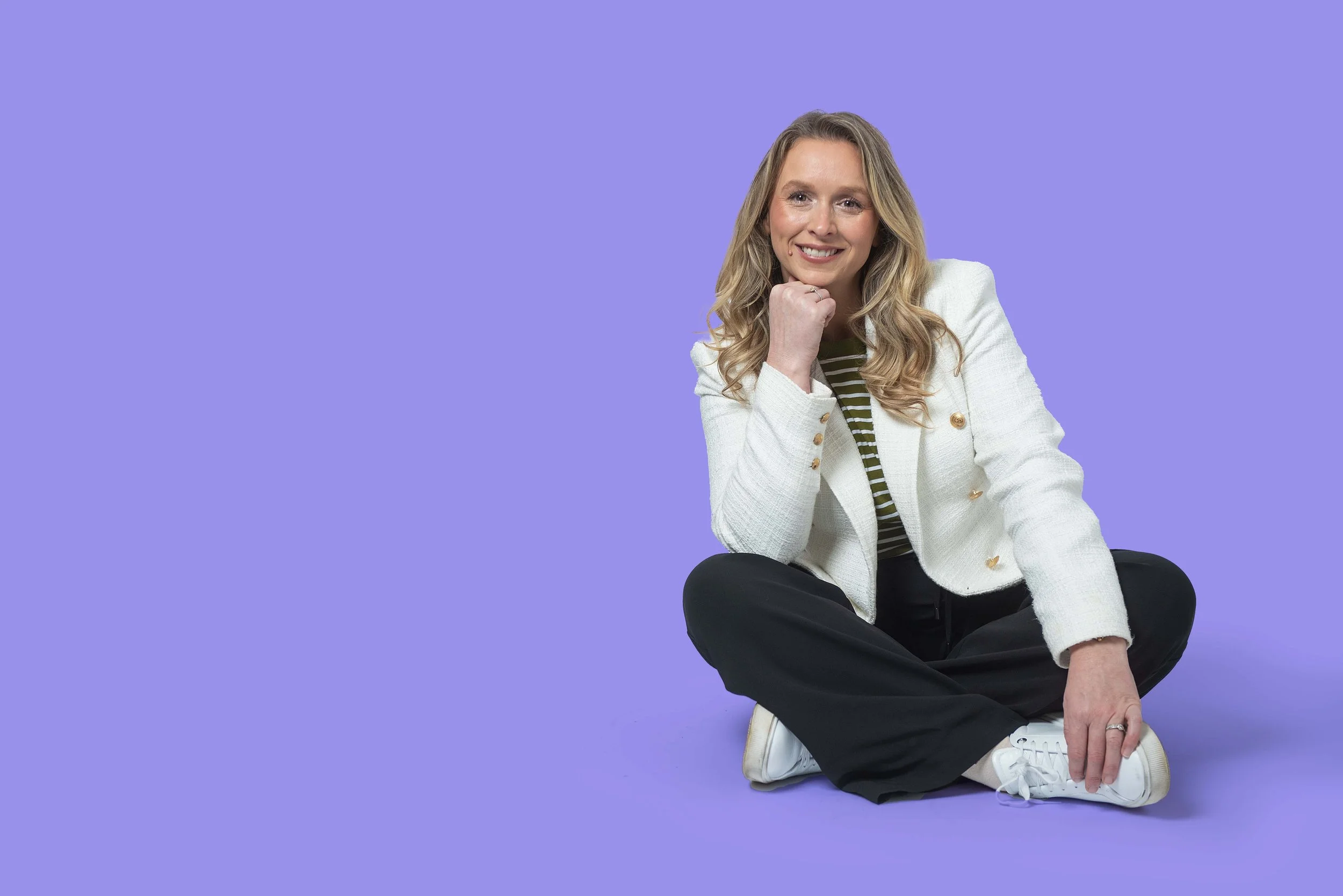 Woman with curly blonde hair in a white blazer sitting cross-legged, lifestyle portrait against a lavender backdrop
