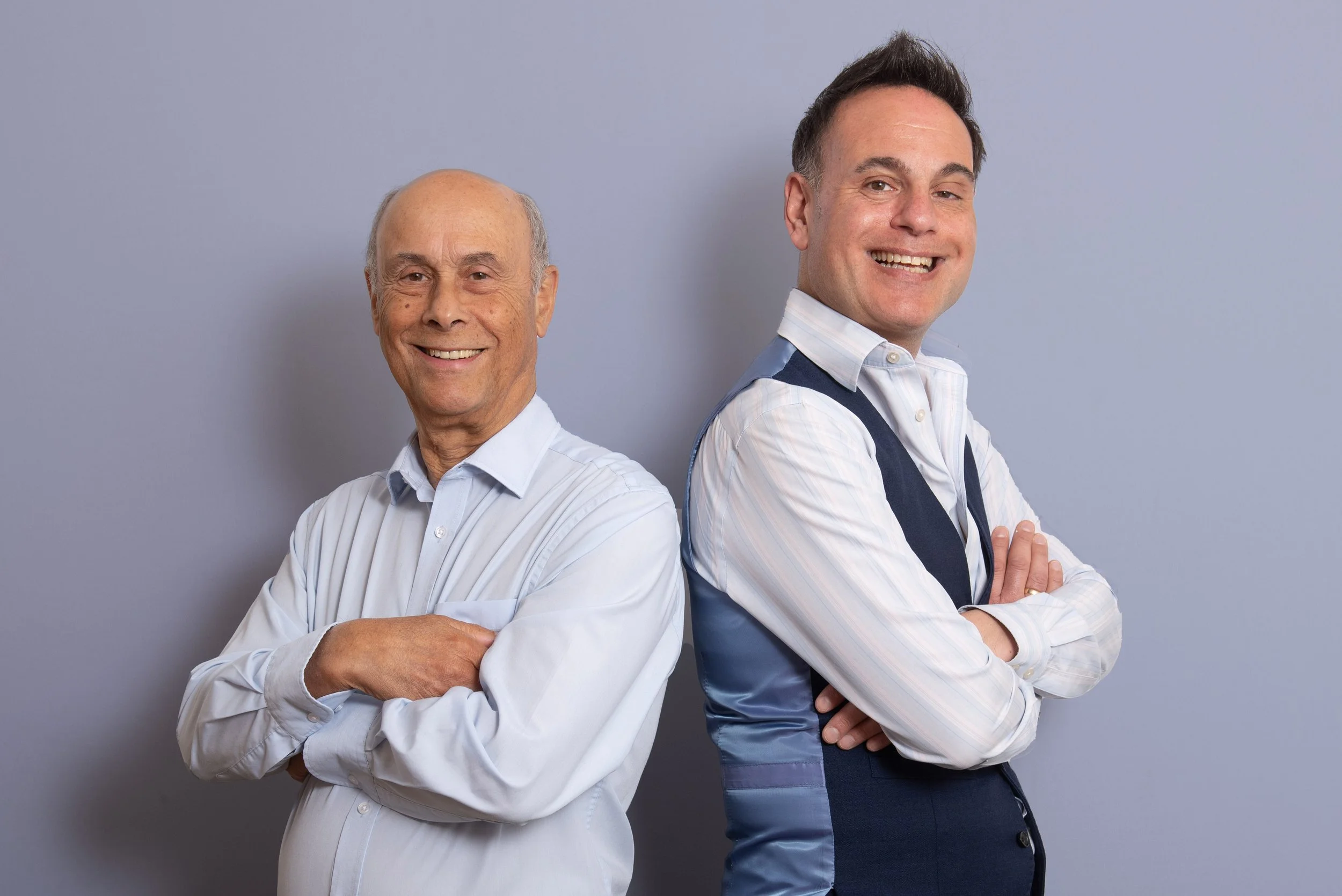 Two smiling businessmen with arms crossed, one older in a light blue shirt and one younger in a navy waistcoat, against a grey studio backdrop. Corporate headshot photography session in Leeds studio